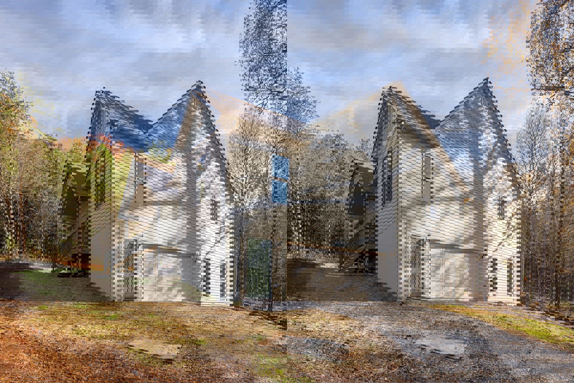 Two-story house with white siding, green front door, attached garage, covered porch, elevated wooden deck, large windows reflecting tree branches, surrounded by grassy lawn and