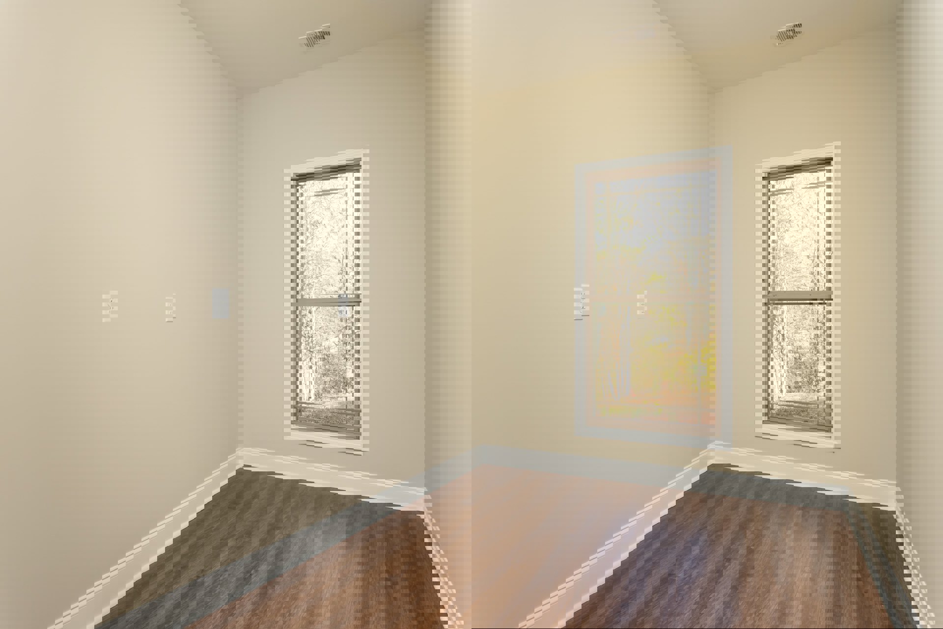 Sunlit room featuring large window with view of green trees, wood laminate flooring, white baseboards, and smooth white ceiling in corner.