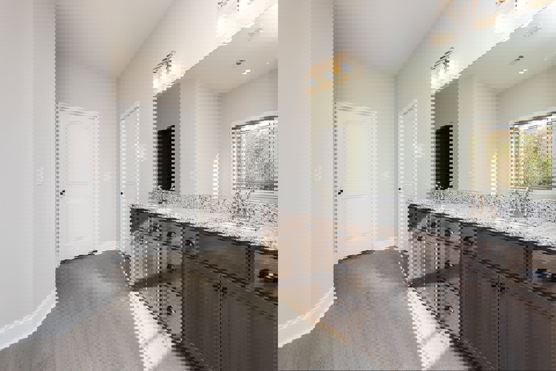 Bathroom with marble countertop double sink vanity, large mirror above, white door with black knob, window showing trees outside, light tile flooring.