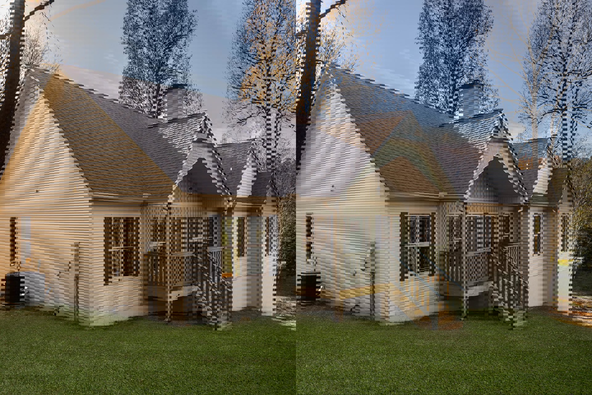 White siding house with covered porch, wooden steps, large windows reflecting trees, black heat pump unit beside exterior, grassy lawn with wooden planter box, mature tree near