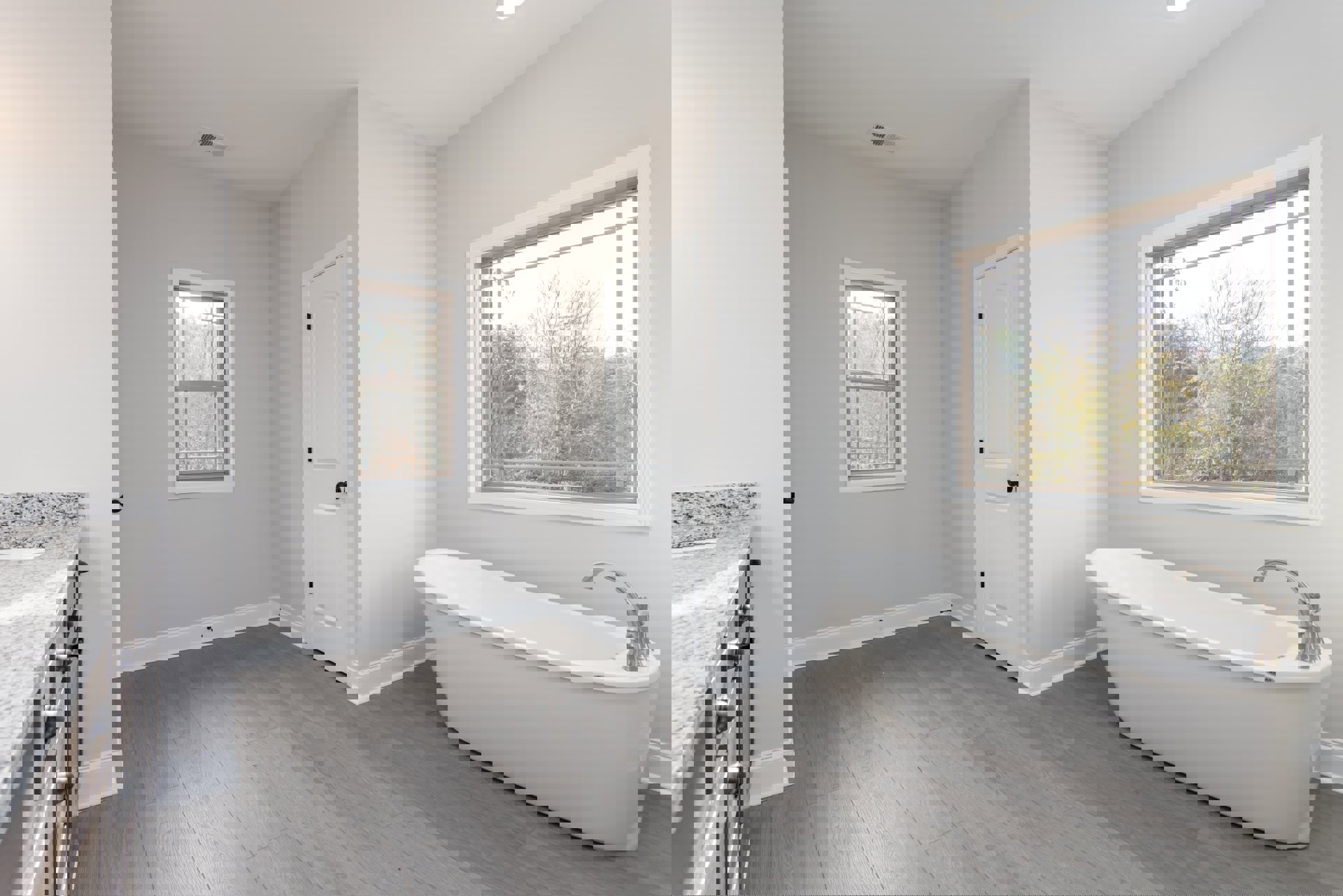 Freestanding white bathtub with chrome faucet beside large windows overlooking trees, wood floor, white paneled door with black knob, light gray tile walls