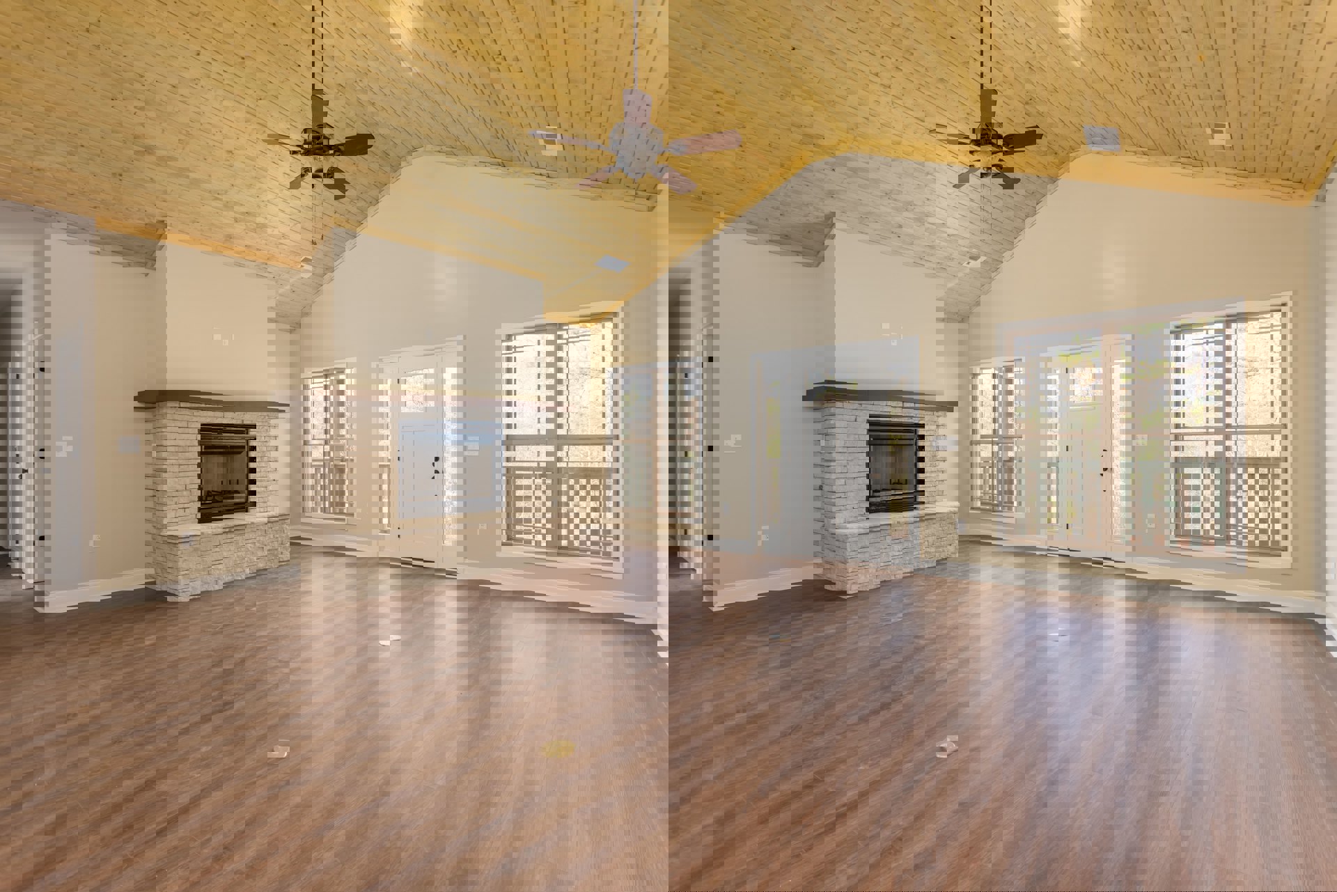 Living room with hardwood floors, white glass-paneled door, stone fireplace with black screen, ceiling fan, large window overlooking deck and trees