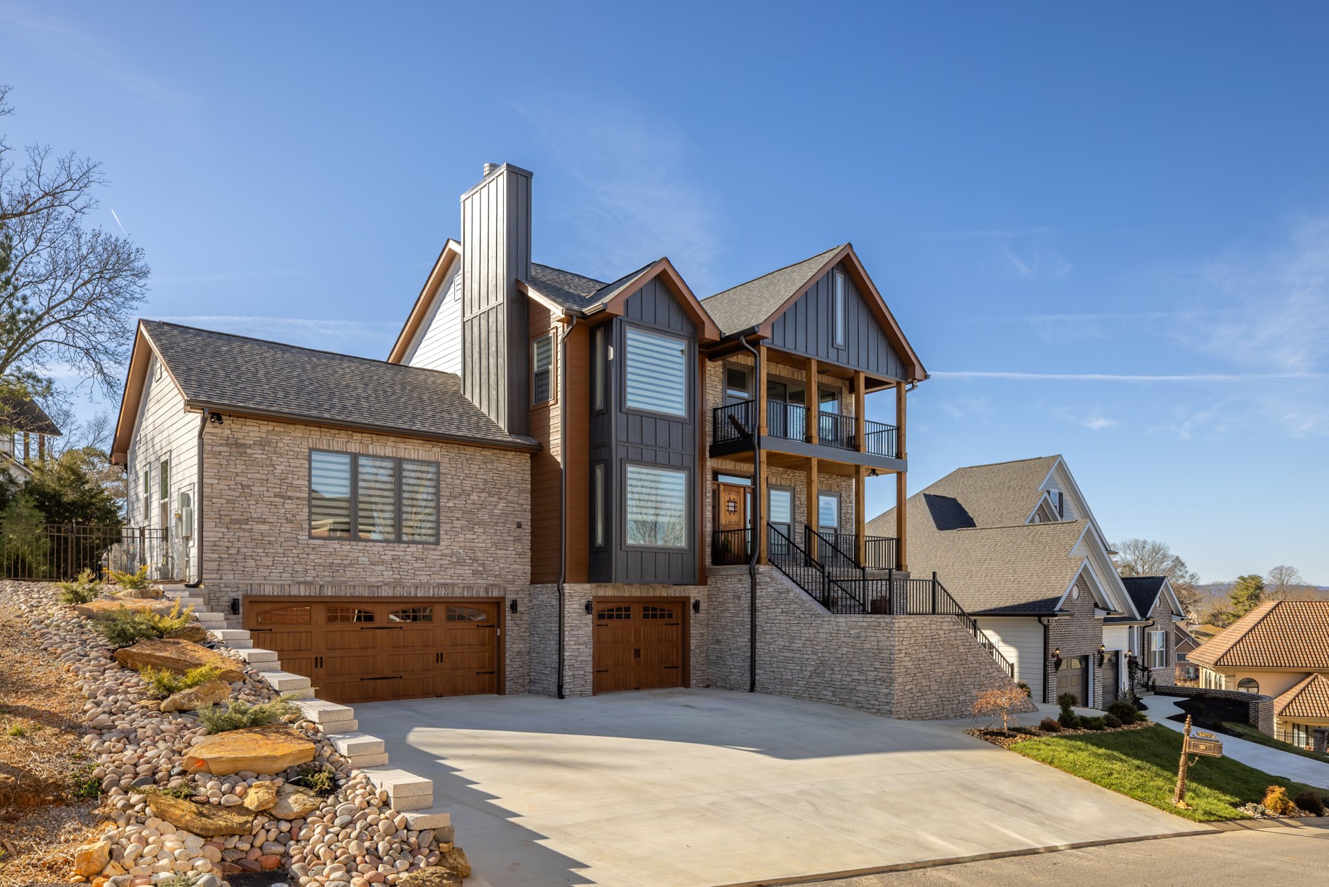 Stone-clad house with attached garage, two driveways, multiple windows including one with blue and white striped blinds, mature tree in front yard, cloudy sky overhead