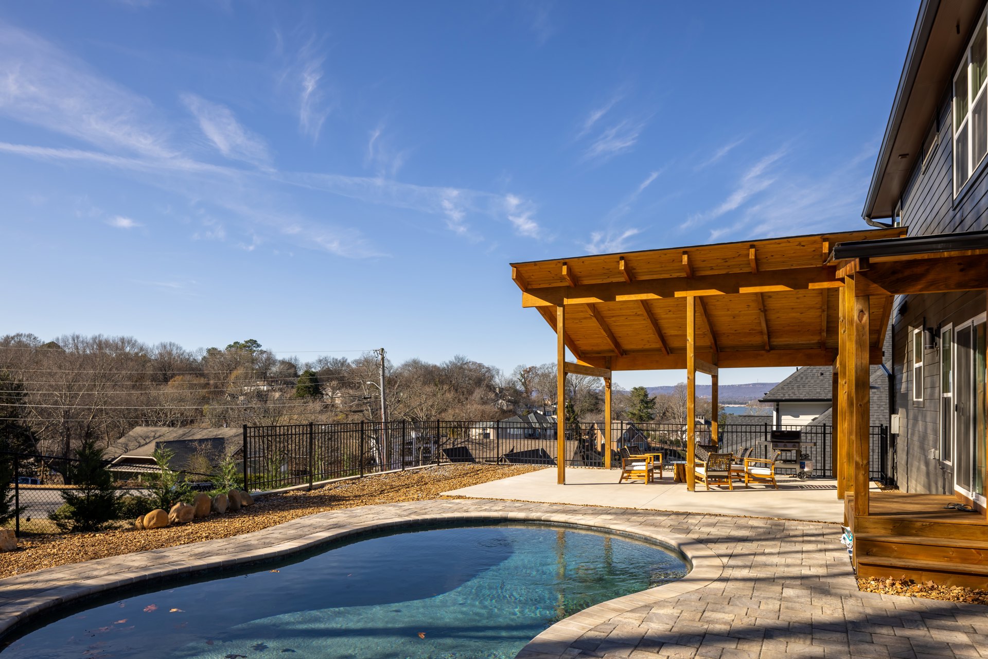 Rectangular swimming pool bordered by stone walkway, covered patio with wooden beams, outdoor lounge chairs, stainless steel grill, modern home exterior, blue sky and scattered