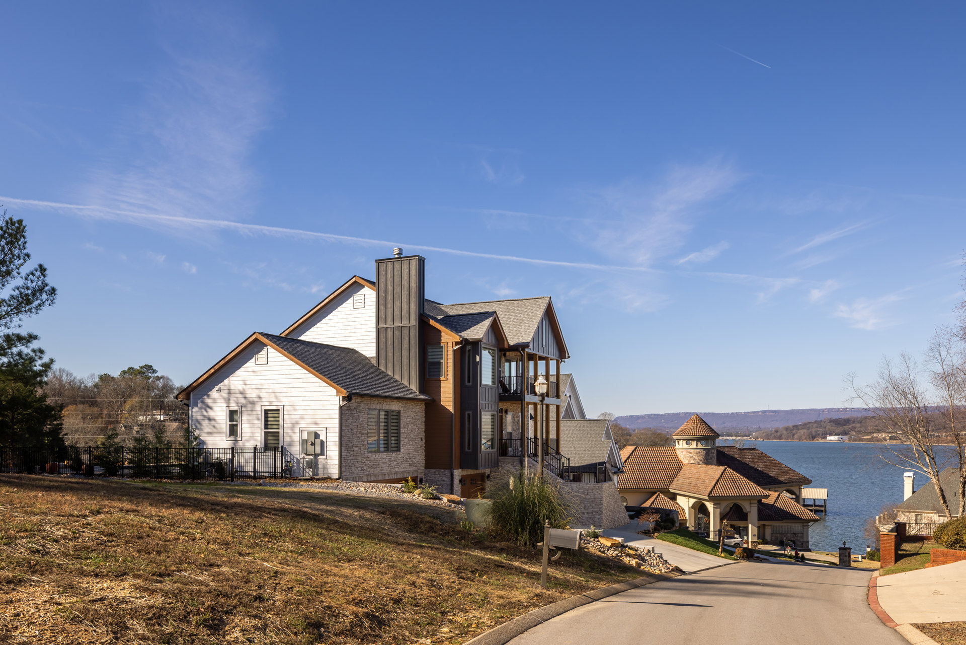 Two-story house with white siding, brown wooden door, white trim, covered porch, brick chimney, metal fence, tree branches overhead, blue sky, and lake in the background