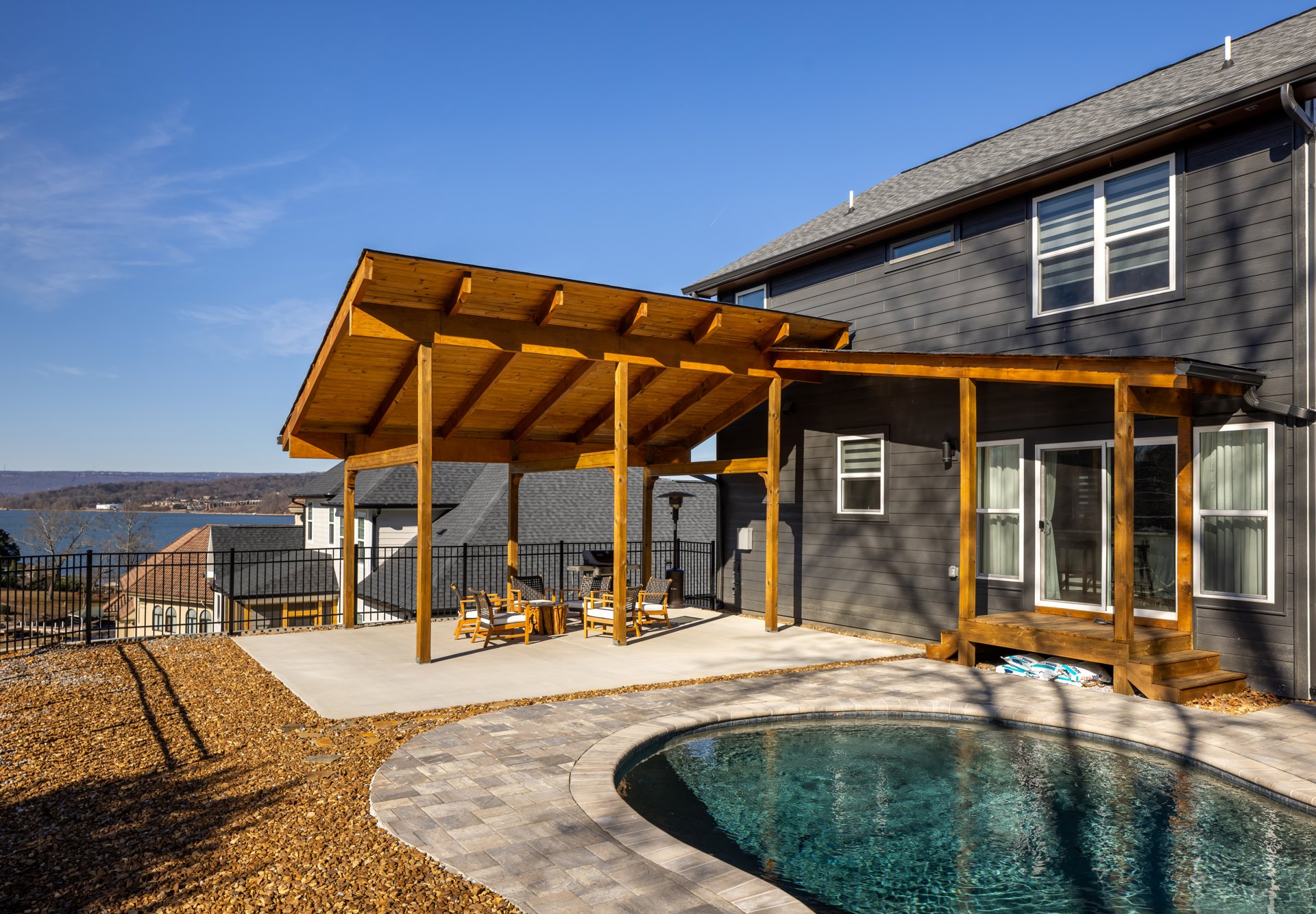 Modern home exterior with stone-edged swimming pool, wooden covered patio furnished with chairs, large white-framed windows, and landscaped outdoor space under a clear sky