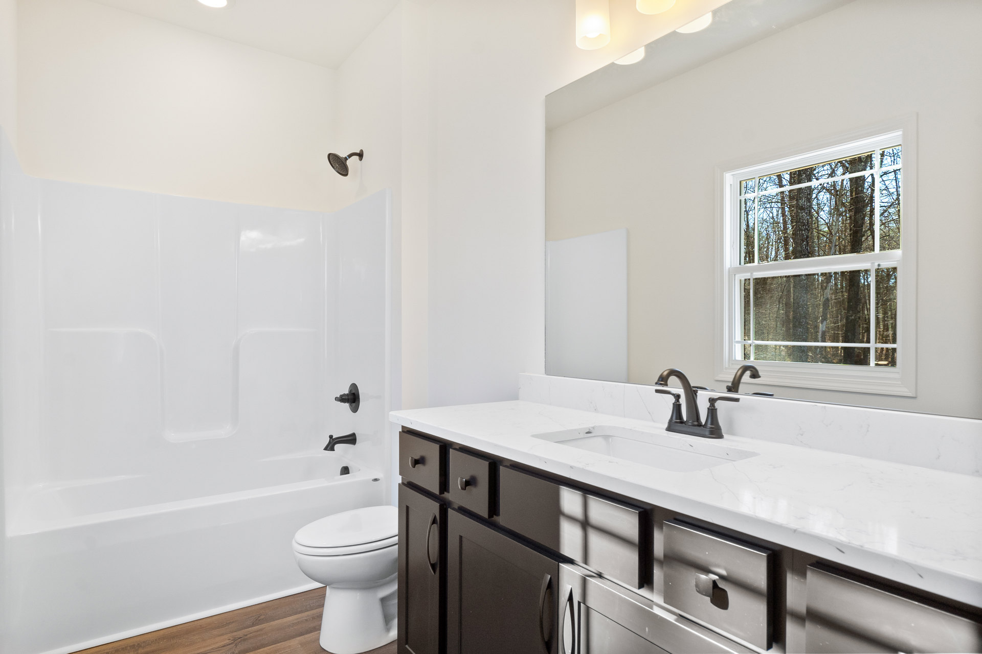 White bathroom featuring a rectangular sink with chrome faucet, white toilet with closed lid, bathtub with wall-mounted shower head, light tile flooring, and window overlooking