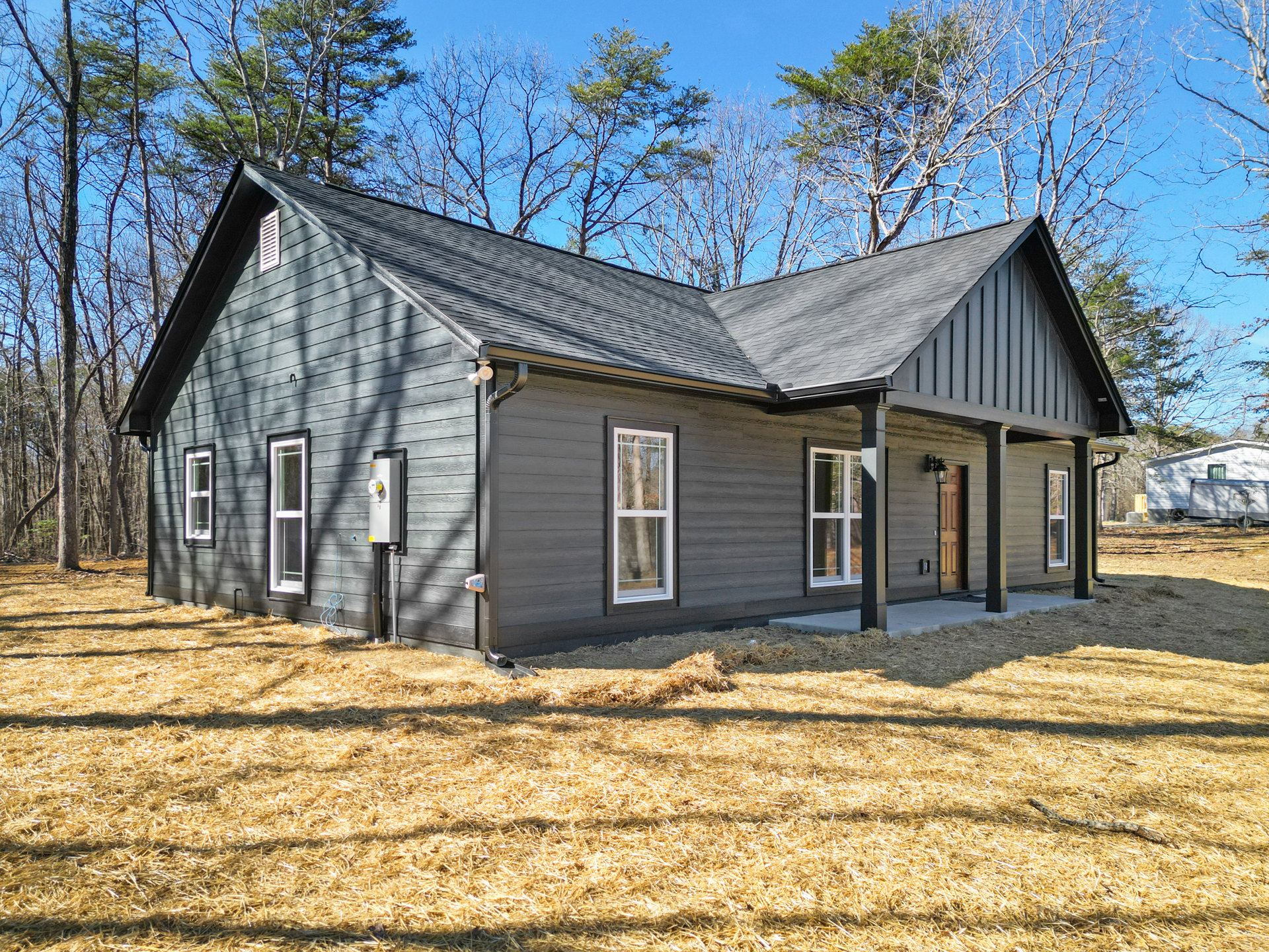Two-story house with multiple large windows, white trim, and a spacious grassy yard featuring mature trees, a parked trailer, and a pile of hay.