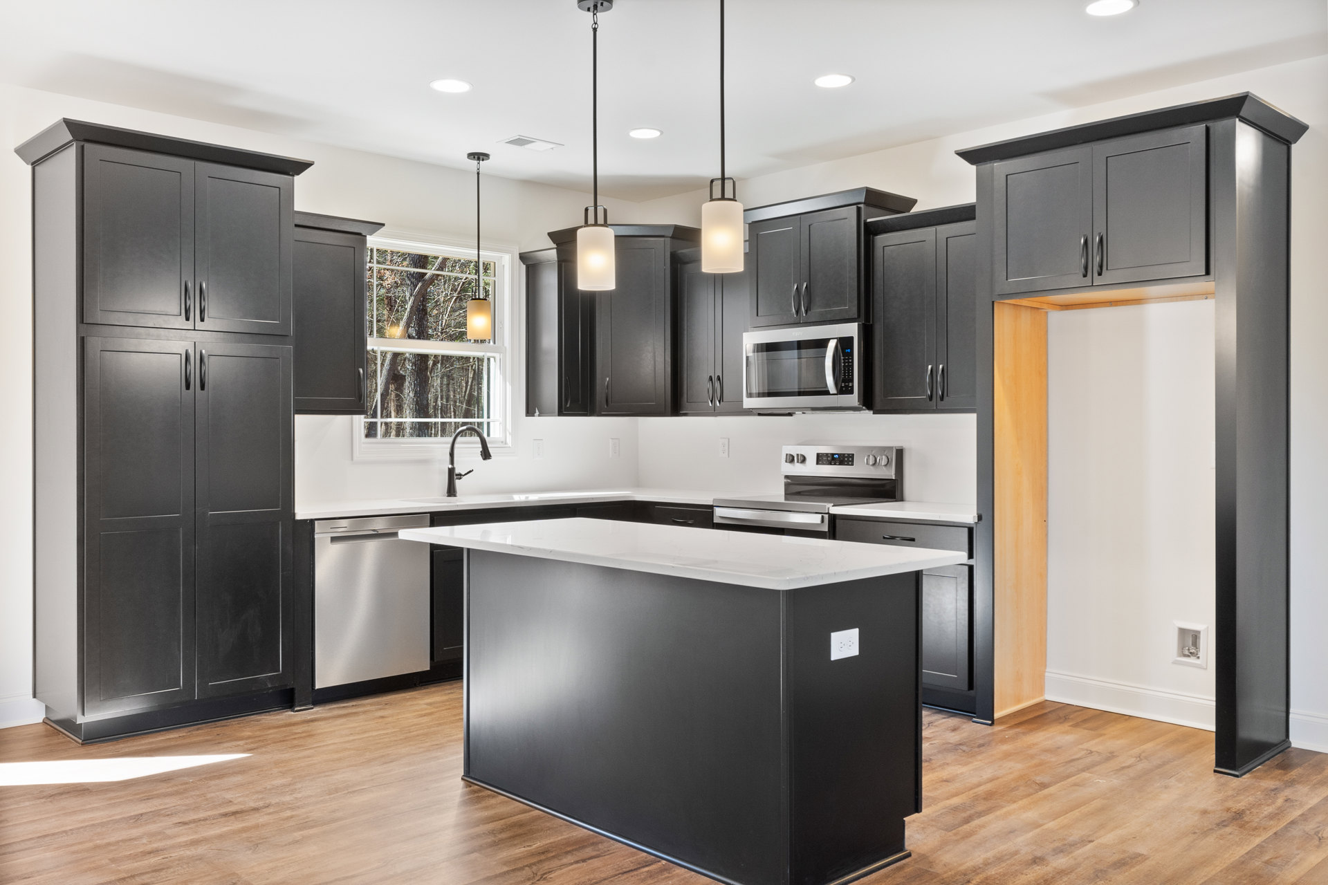 Black kitchen cabinets paired with a white island, white countertops, stainless steel microwave, black cabinet handles, modern pendant lighting, and light wood flooring.