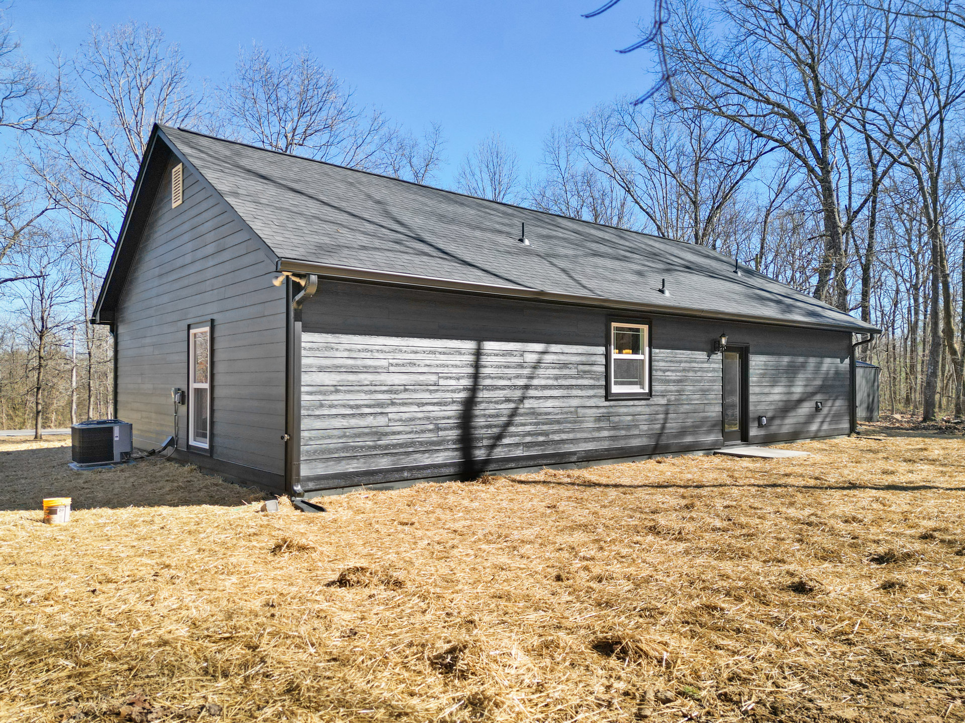 Grey siding house with white-trimmed window, gutter, and security camera, surrounded by grassy yard, mature trees, and metal utility box