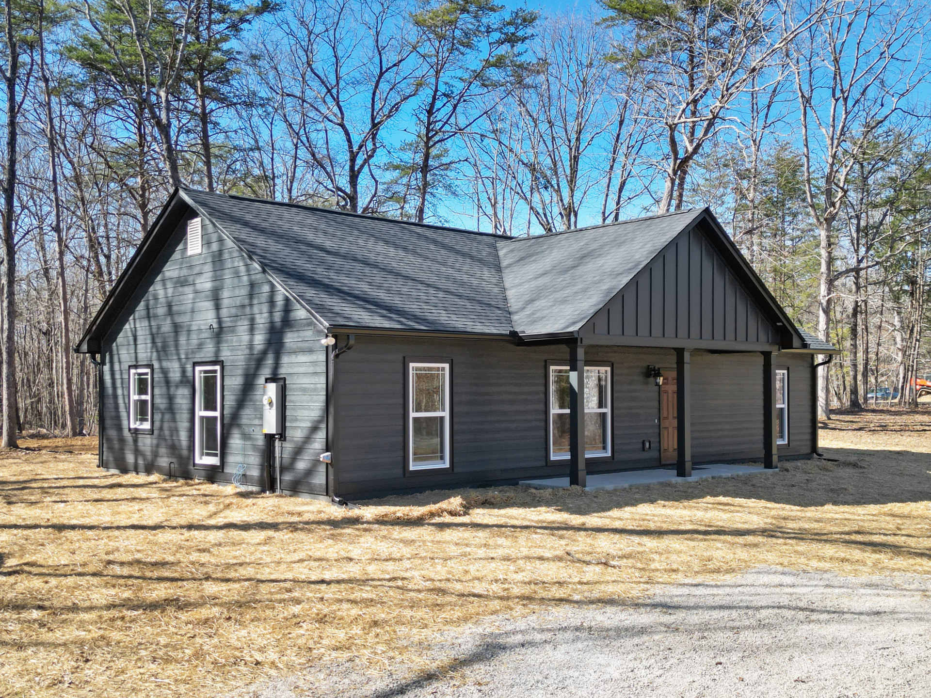 Two-story house with black roof, white-framed windows, light siding, and a pile of hay in front; surrounded by tall trees in the background