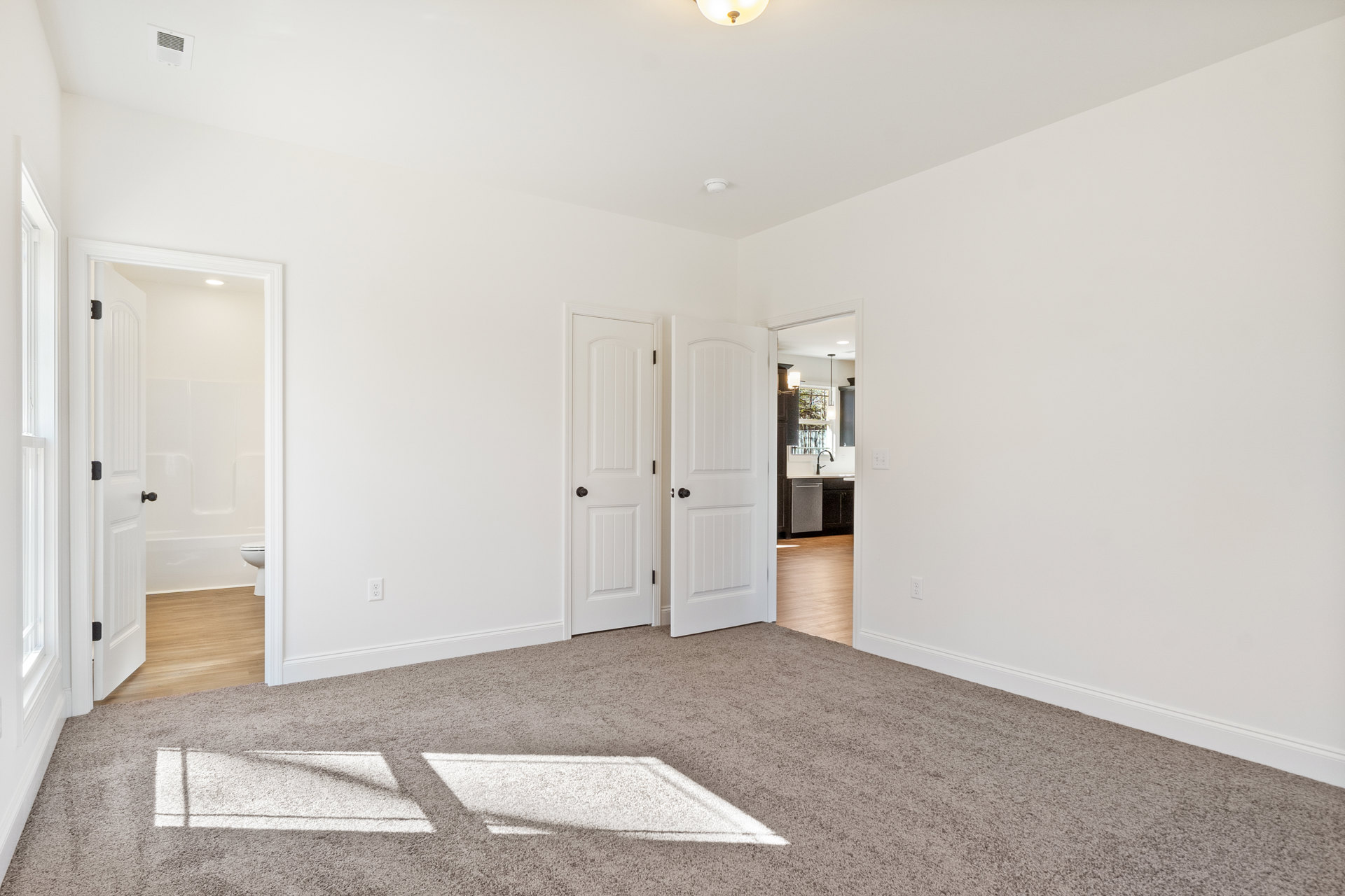 White-walled room with light carpet flooring, white door featuring a black knob, wall vent, and partial view of a kitchen sink beneath a hanging light fixture