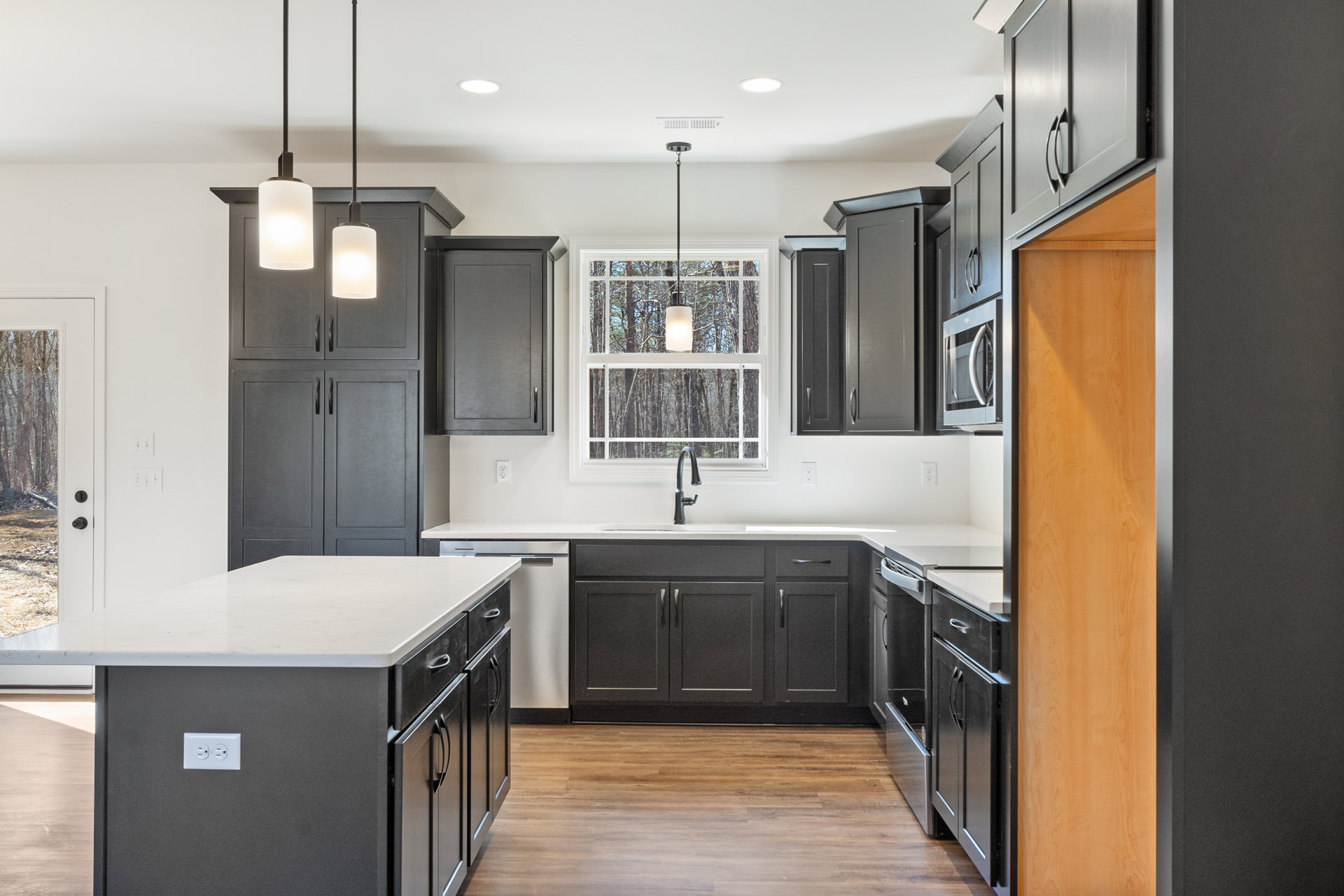 Black kitchen cabinets with white countertops, central island with white surface, stainless steel fixtures, rectangular pendant light, sunlight streaming through window onto
