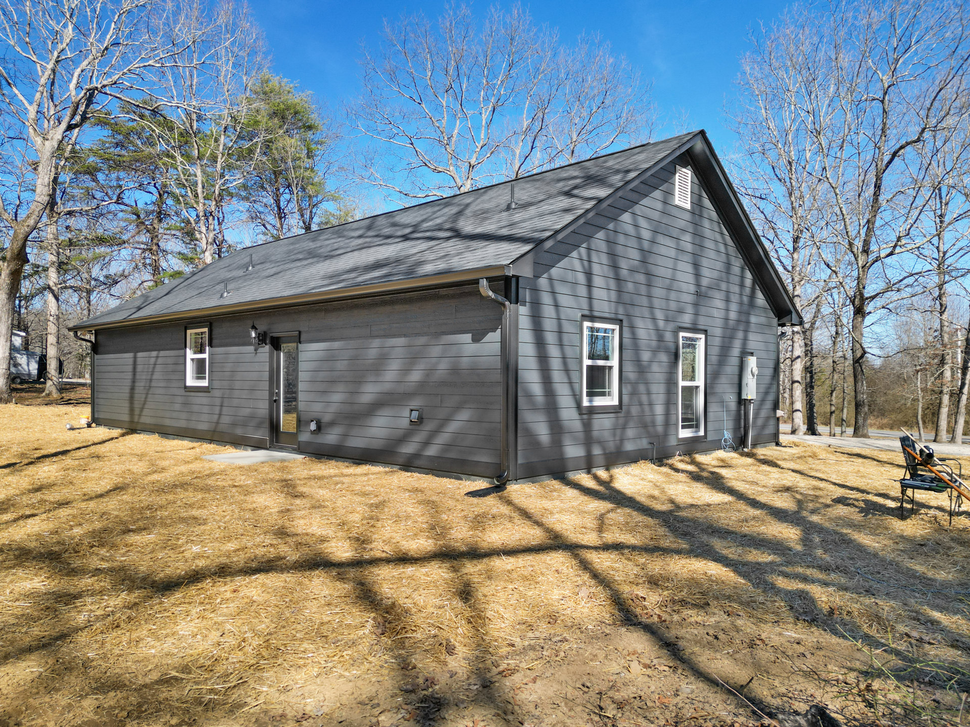 Grey cottage-style home with white-framed windows, front door, and outdoor chair with paddles, surrounded by trees and tree shadows on the lawn
