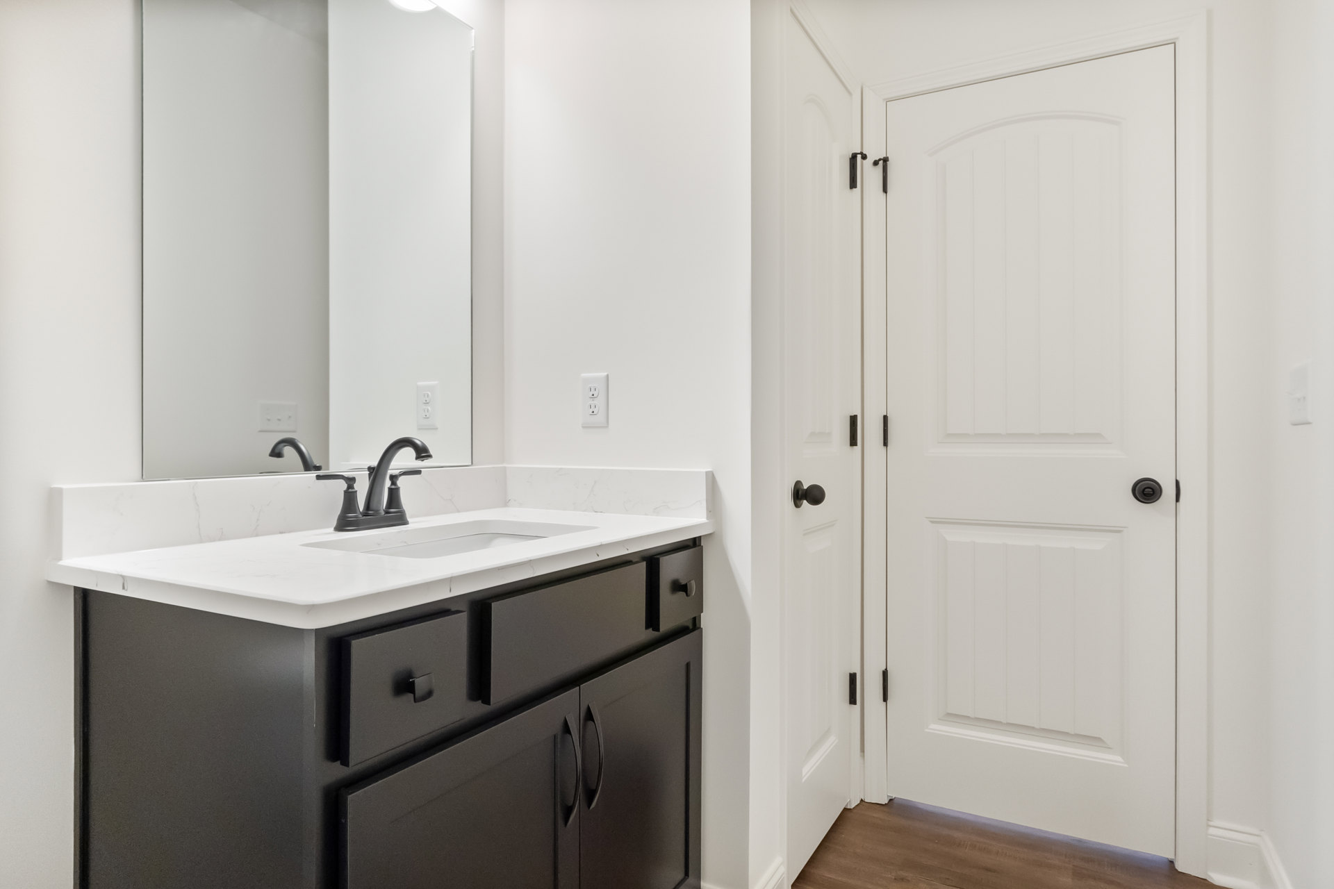 White bathroom door with black handle, black framed mirror above black faucet and sink set in white countertop, light tile walls and cabinetry.