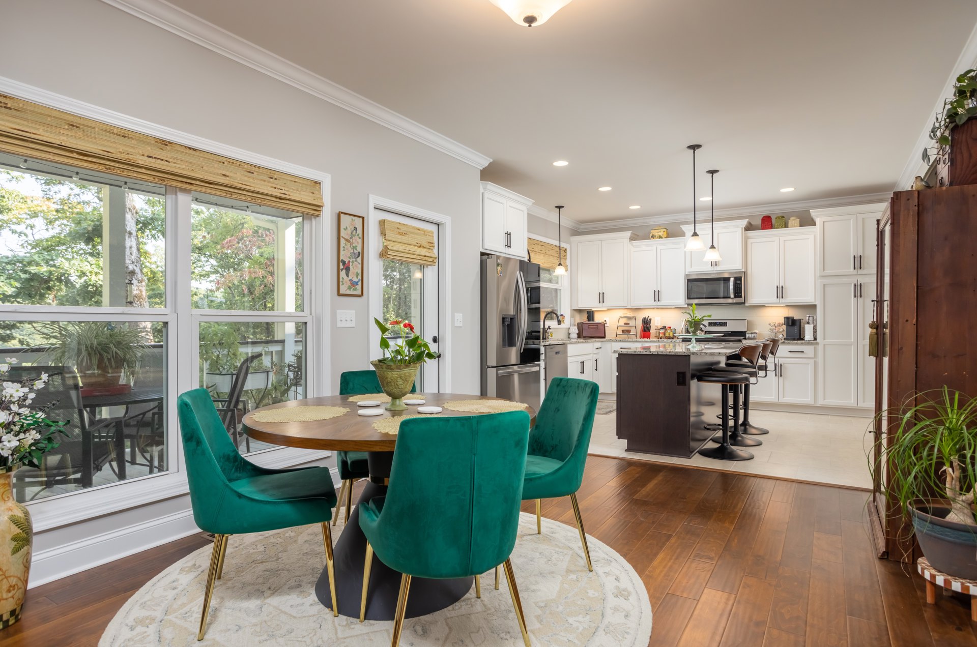 Dining area with green chairs and gold legs around a wooden table, white kitchen cabinets in the background, potted plant and vase on the table, microwave built into cabinetry