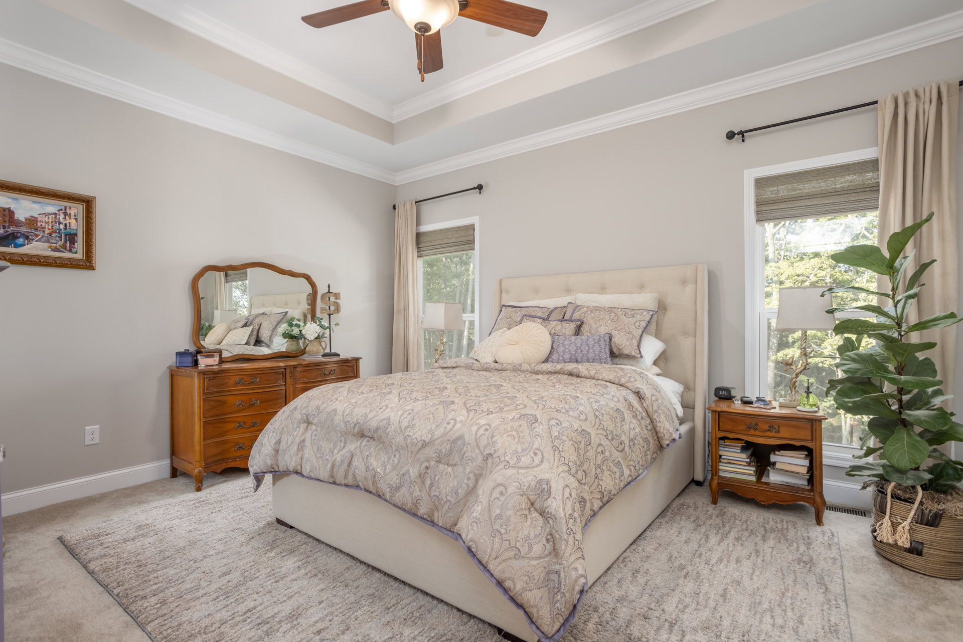 Bedroom with a bed featuring white and purple comforter, pillows, wooden dresser, nightstand holding books and a phone, potted plant, framed picture of a bridge over a river on the
