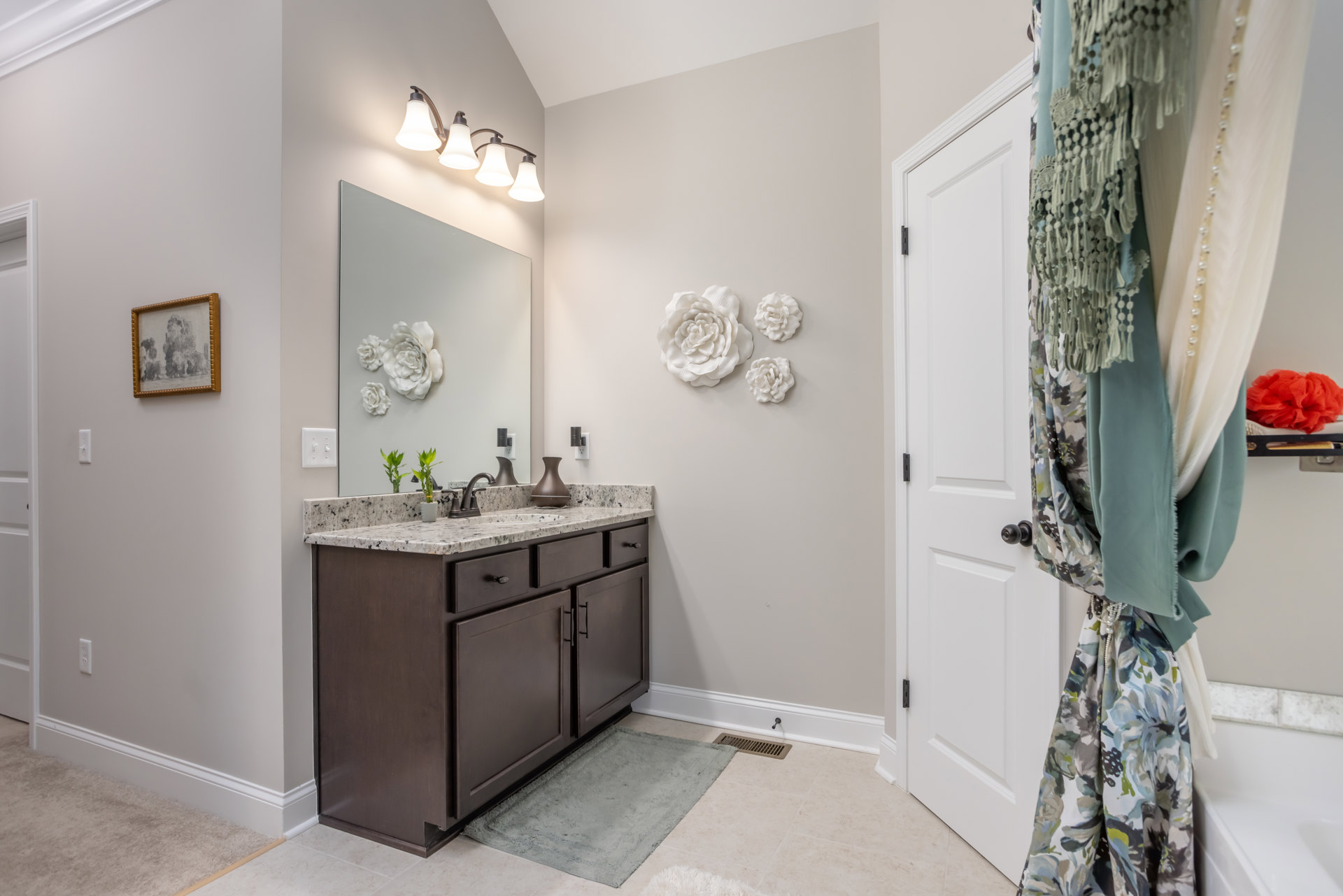 White bathroom with rectangular mirror above a stone countertop, undermount sink, chrome faucet, brown vase, framed tree artwork, wall-mounted row of lights, and light tile