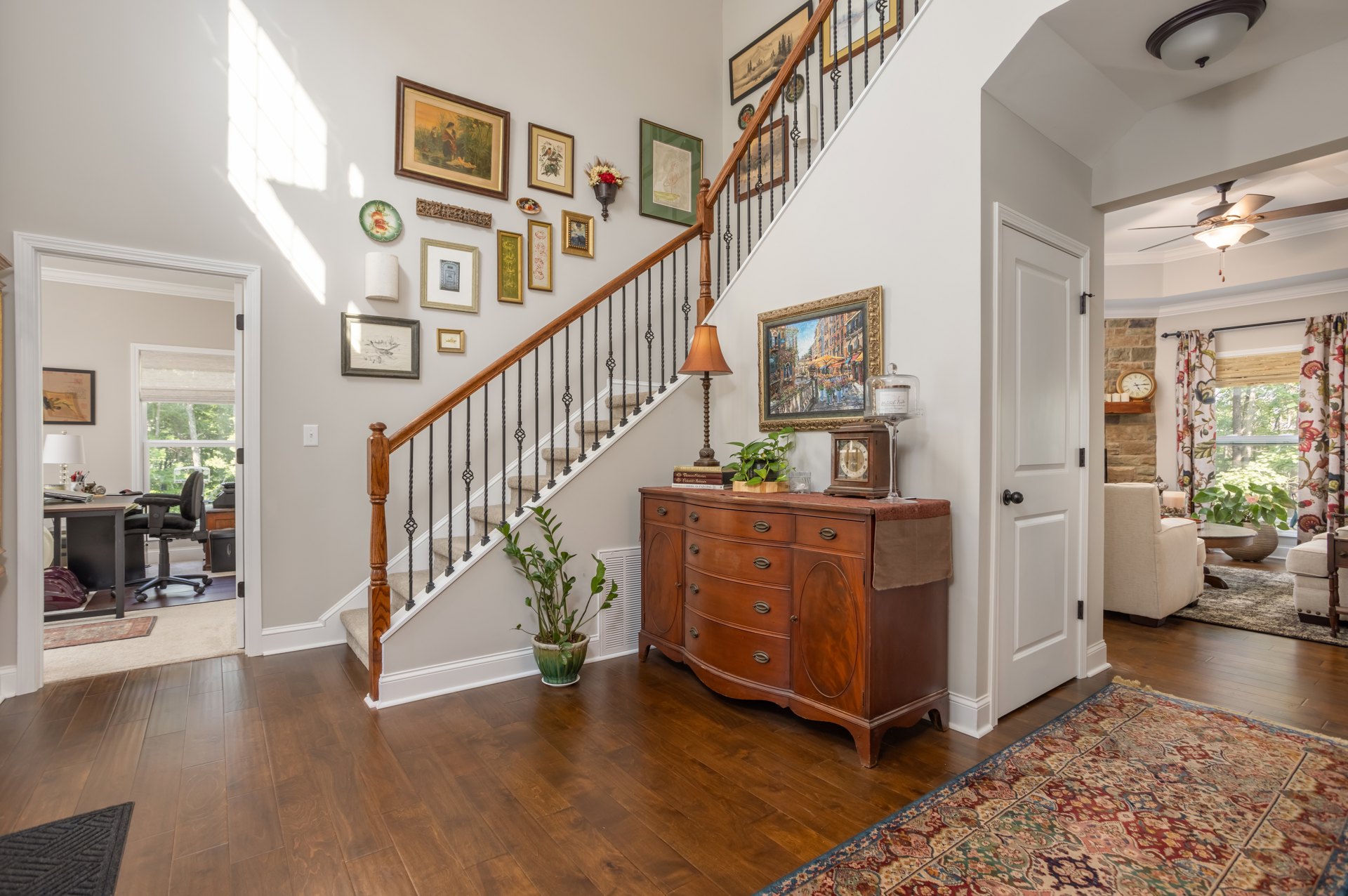 Wood staircase with white risers, patterned rug on laminate flooring, dresser topped with decorative plate and framed photos, potted plant beside dresser, wall adorned with framed