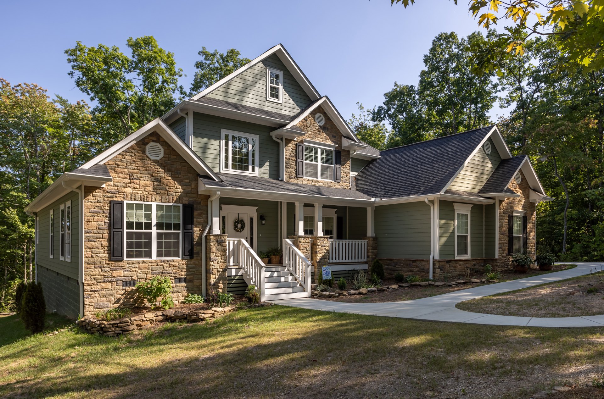 Two-story home with light siding, white-framed windows, covered porch, paved driveway, potted plant near entrance, mature trees in background