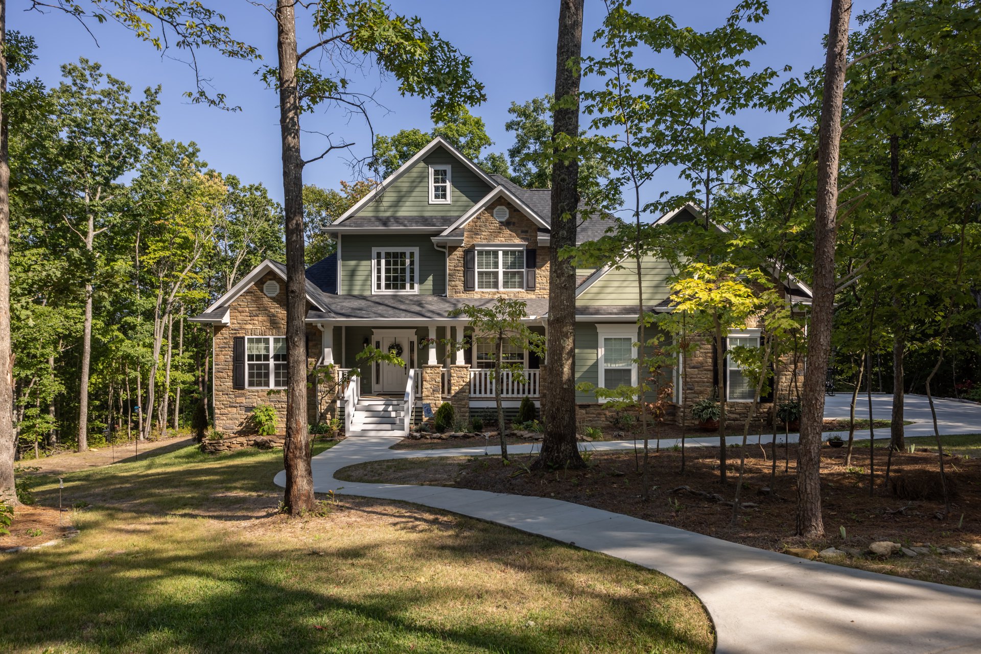 Two-story home surrounded by mature trees, white-framed windows with blinds, covered porch, manicured grass lawn, potted plant near entrance, sidewalk bordering the property.