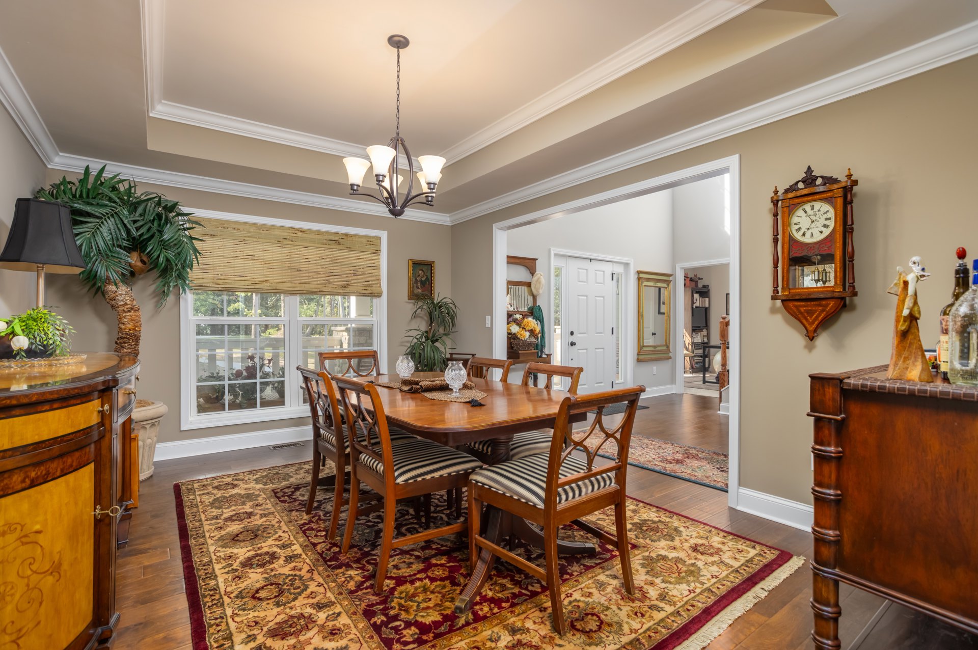 Dining room with wooden table and upholstered chairs on patterned rug, large wall clock with roman numerals, light cabinetry, neutral walls, and recessed ceiling lighting