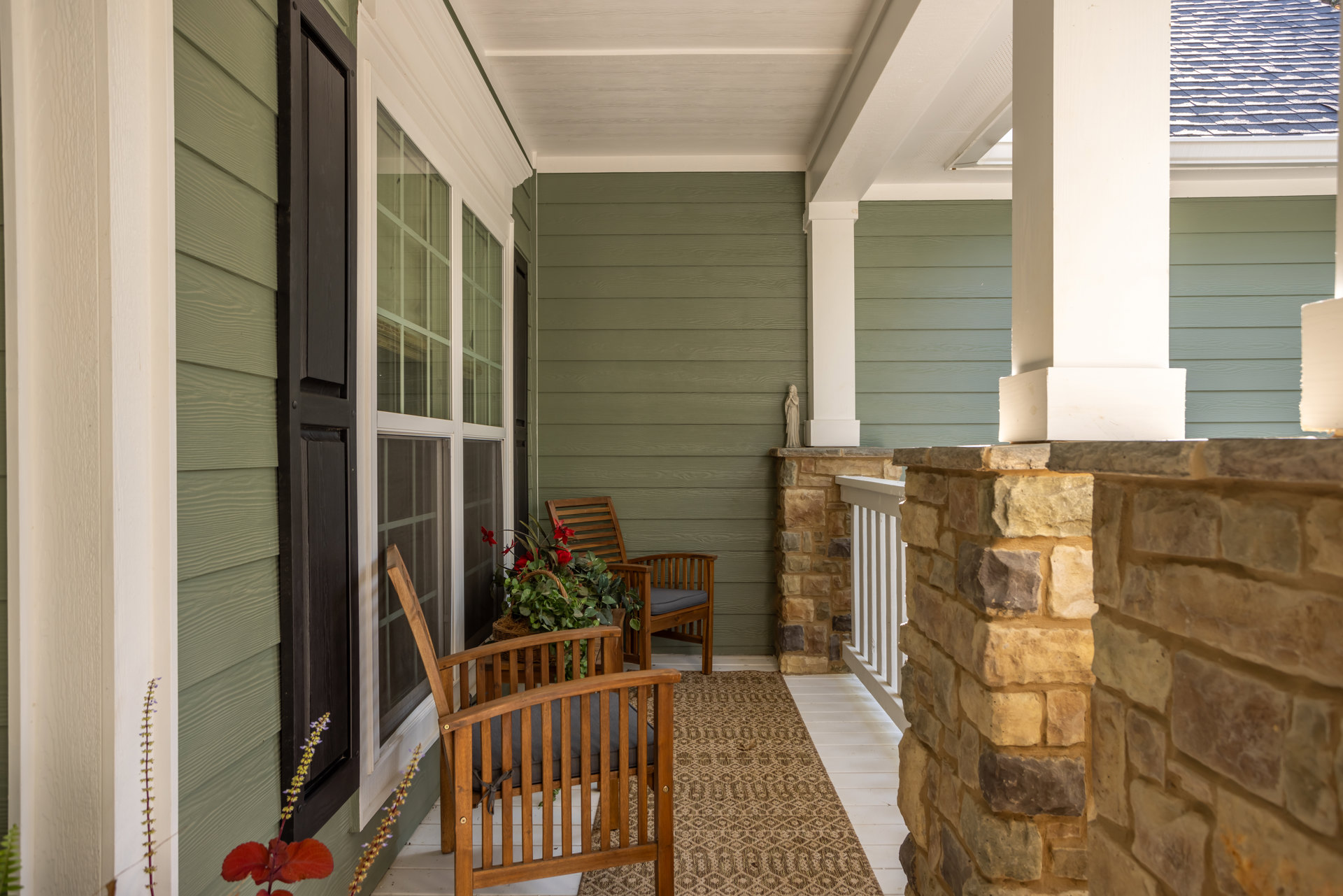 Covered porch with wooden chairs, patterned rug, basket of flowers, brick wall, and white handrail