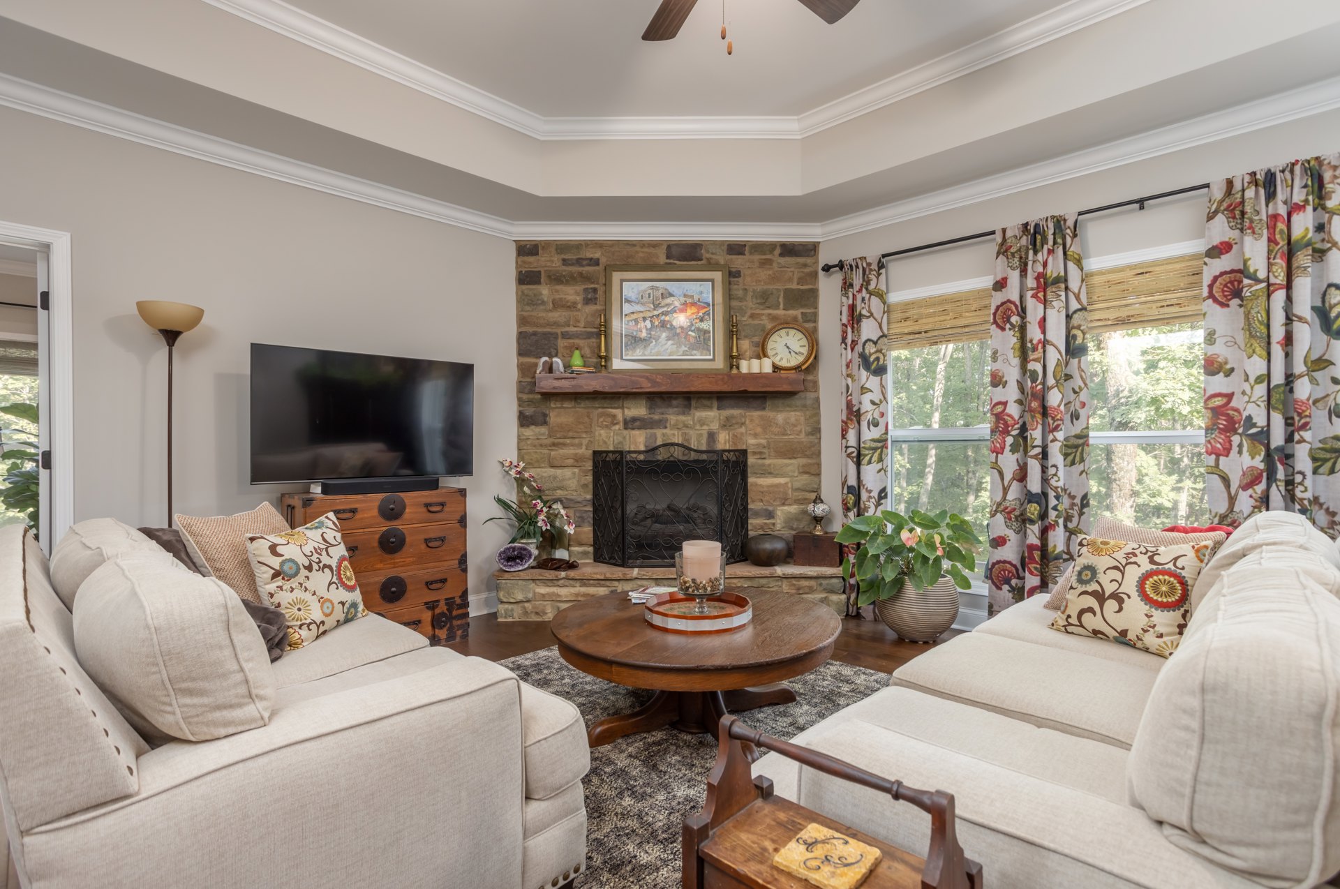 Living room with stone fireplace, two gray couches with patterned pillows, wooden coffee table, TV on a stand, framed market artwork on wall, potted plant, dresser, and glass