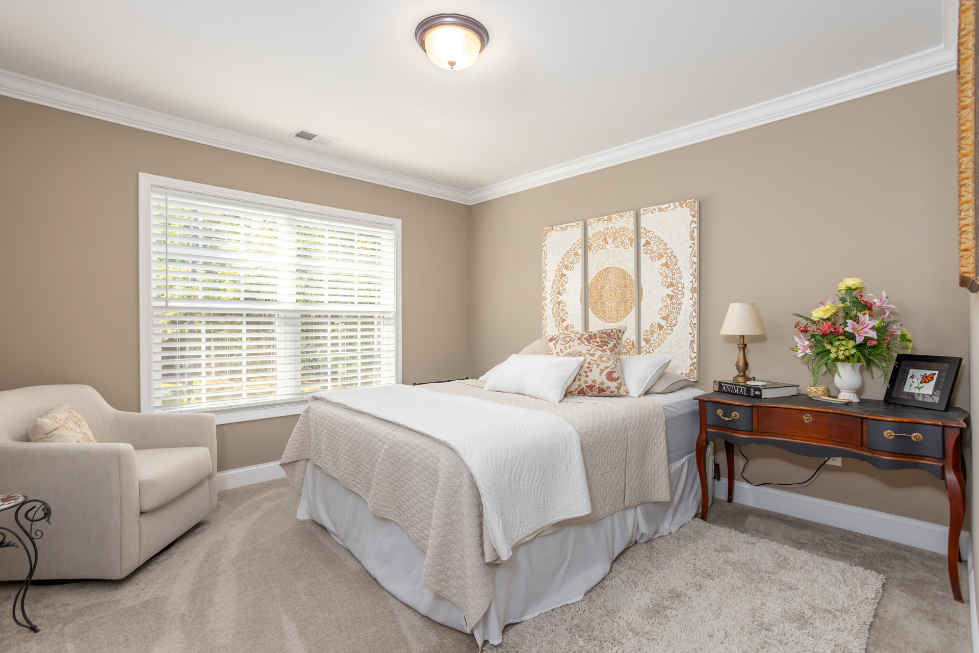 Bedroom with upholstered bed topped with pillows, small side table holding a vase of flowers, white couch, framed butterfly and floral artwork on wall, window with white blinds