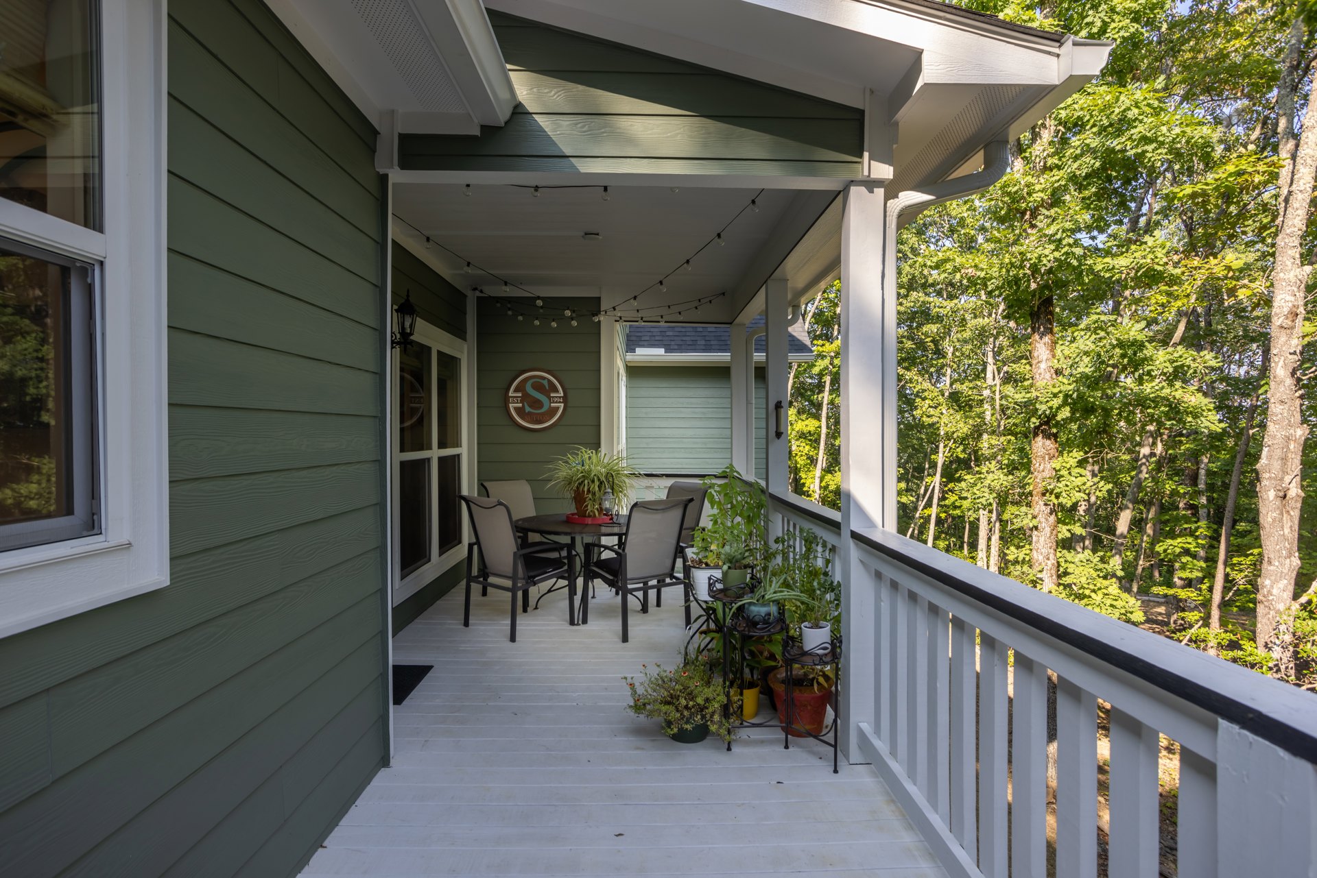 Covered porch with white siding, wooden chairs and tables, potted plant, handrail, and leafy trees in background