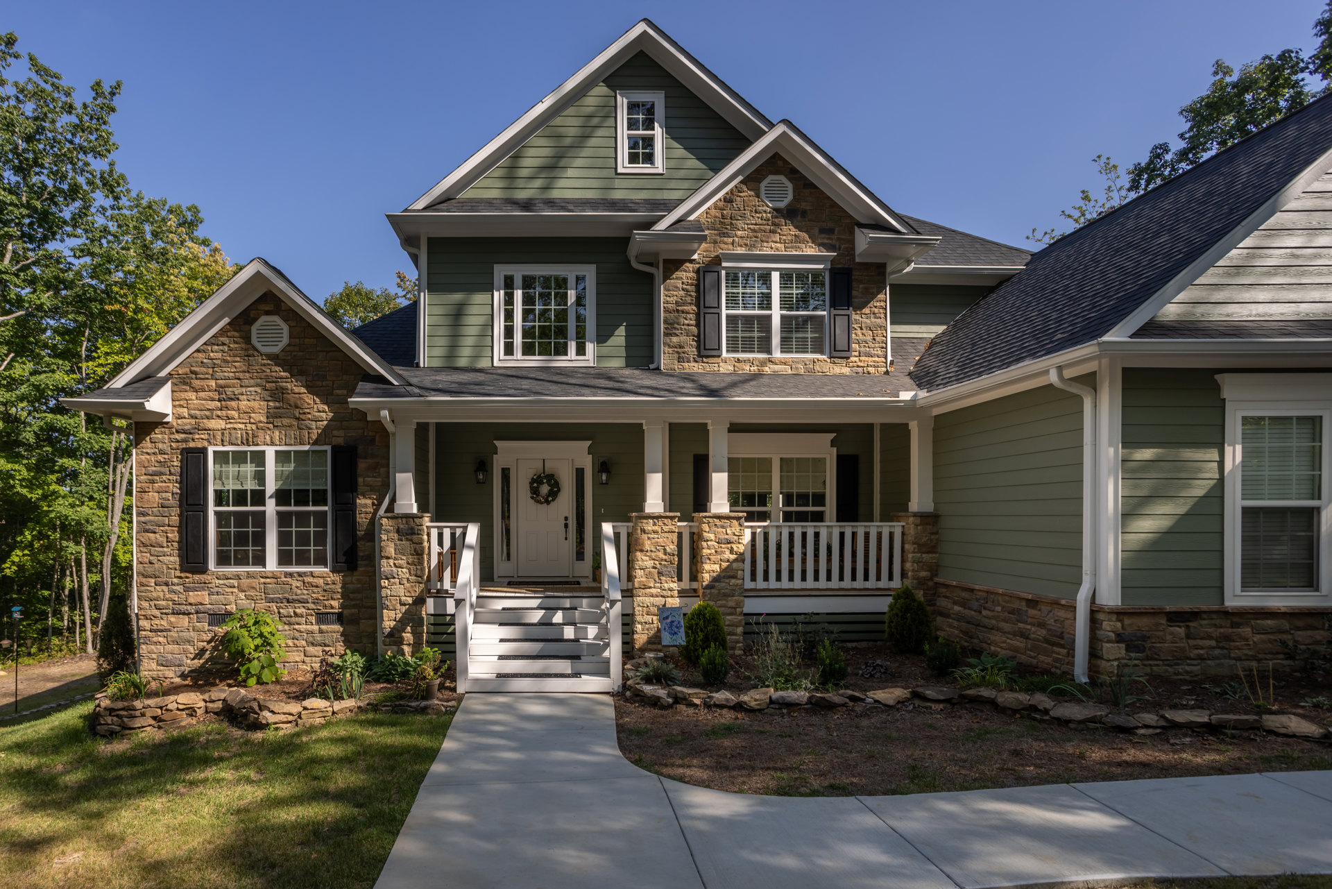 Two-story house with stone and siding exterior, white-trimmed windows, white front door with wreath, green lawn, driveway, and bushes along the stone wall