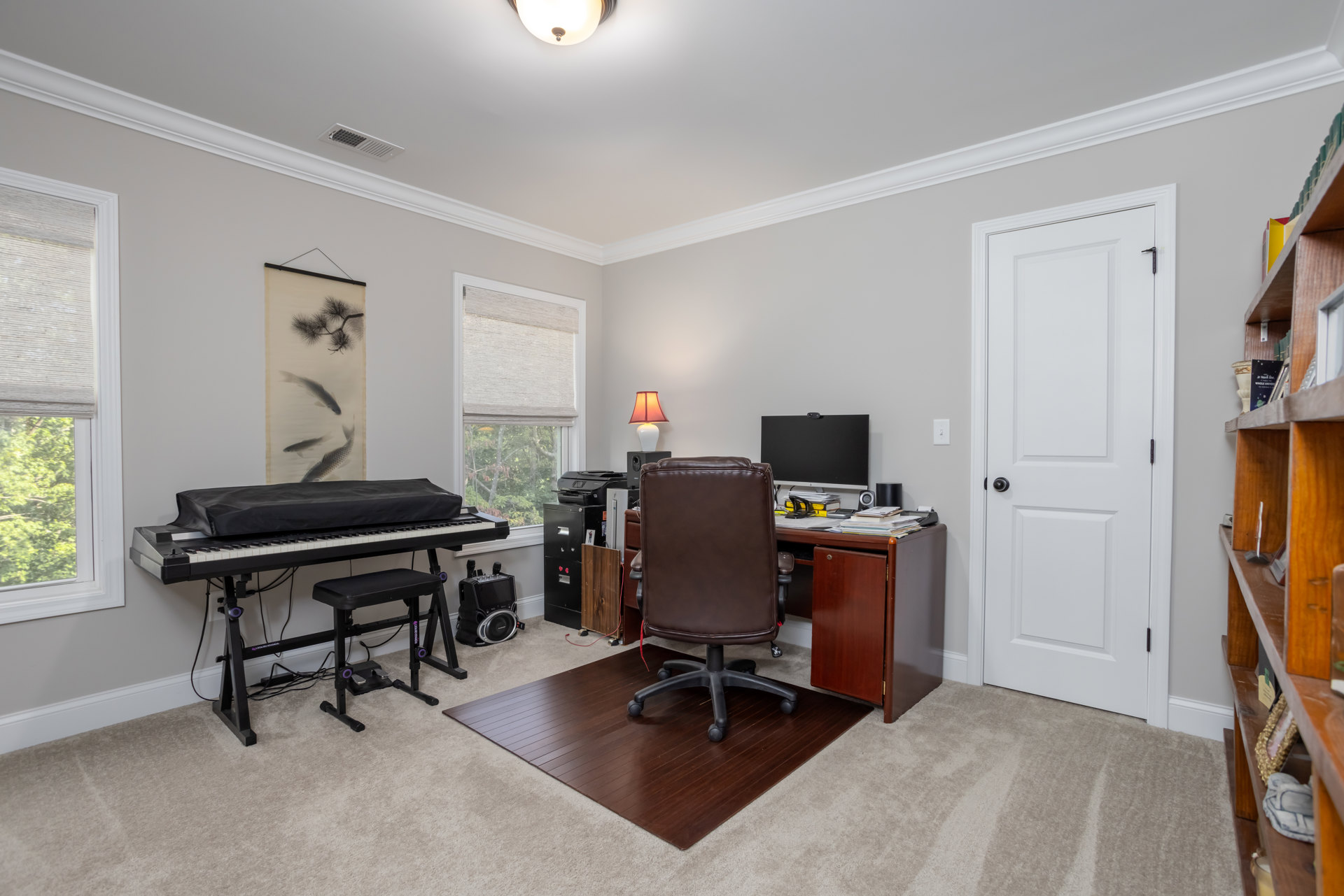 Home office with brown leather chair at wood desk, computer monitor, lamp with red shade, white door with black knob, piano covered in black fabric, black chair with purple circle