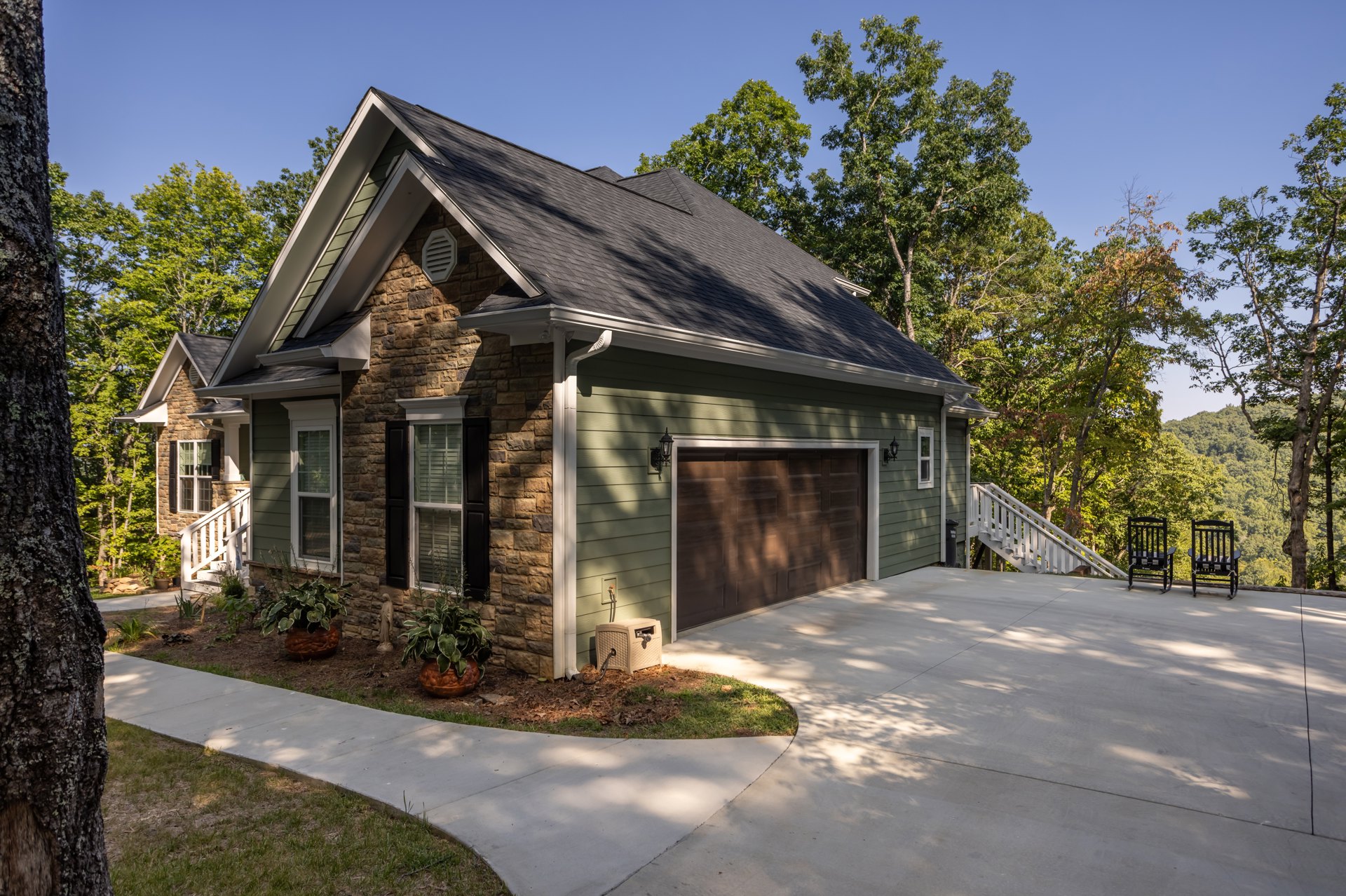 Modern home with gray siding, attached garage, concrete driveway, mature trees, potted plant near entrance, plastic hose reel, outdoor baby crib, chair secured to railing