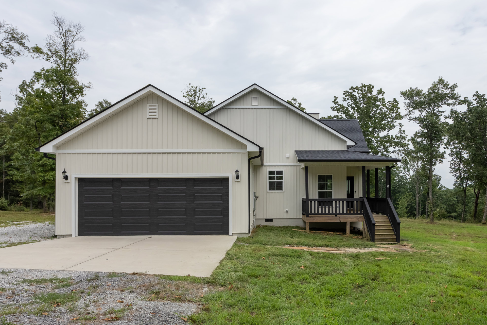 White house with black-trimmed garage door, covered porch, wooden deck with black railing, concrete driveway bordered by grass and rocks, American Gothic House visible in