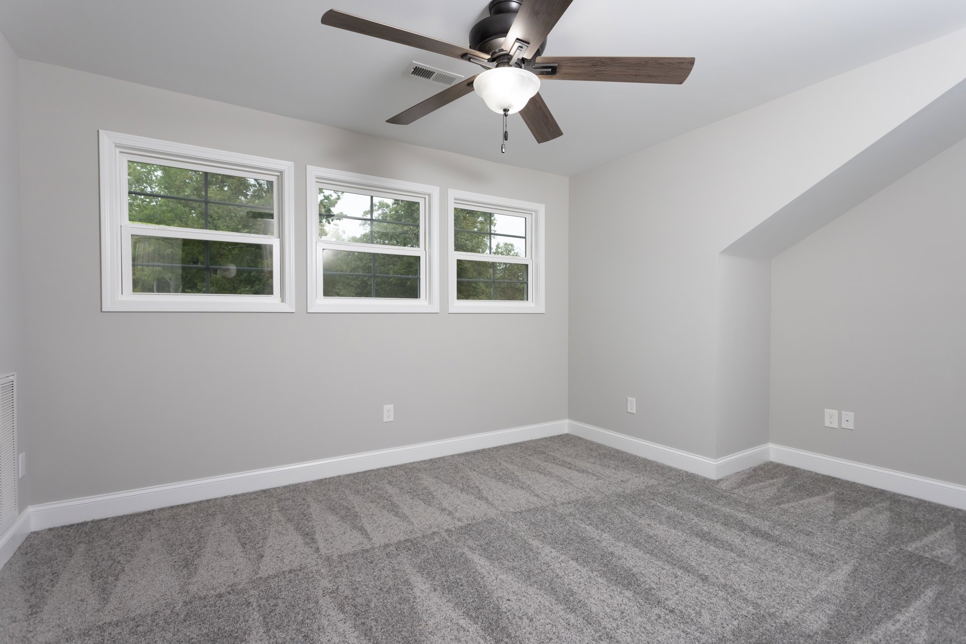 Ceiling fan with light fixture centered in a room featuring grey carpet, white-framed windows, and smooth plaster walls.