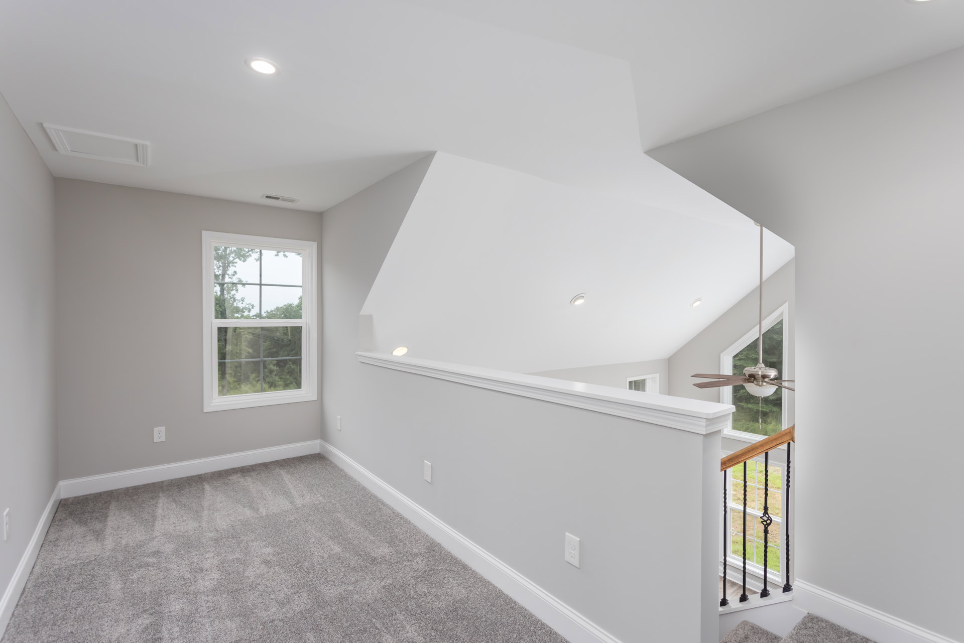 White-walled room featuring a carpeted floor, wooden staircase with metal railing, large window overlooking trees, and ceiling fan.