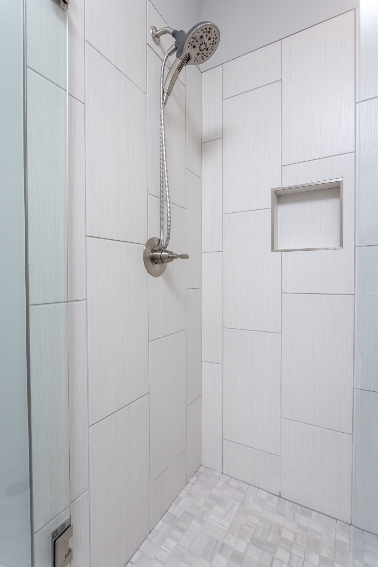 White tile shower with chrome handheld shower head, hose, and square faucet on tiled wall; partial view of white door.
