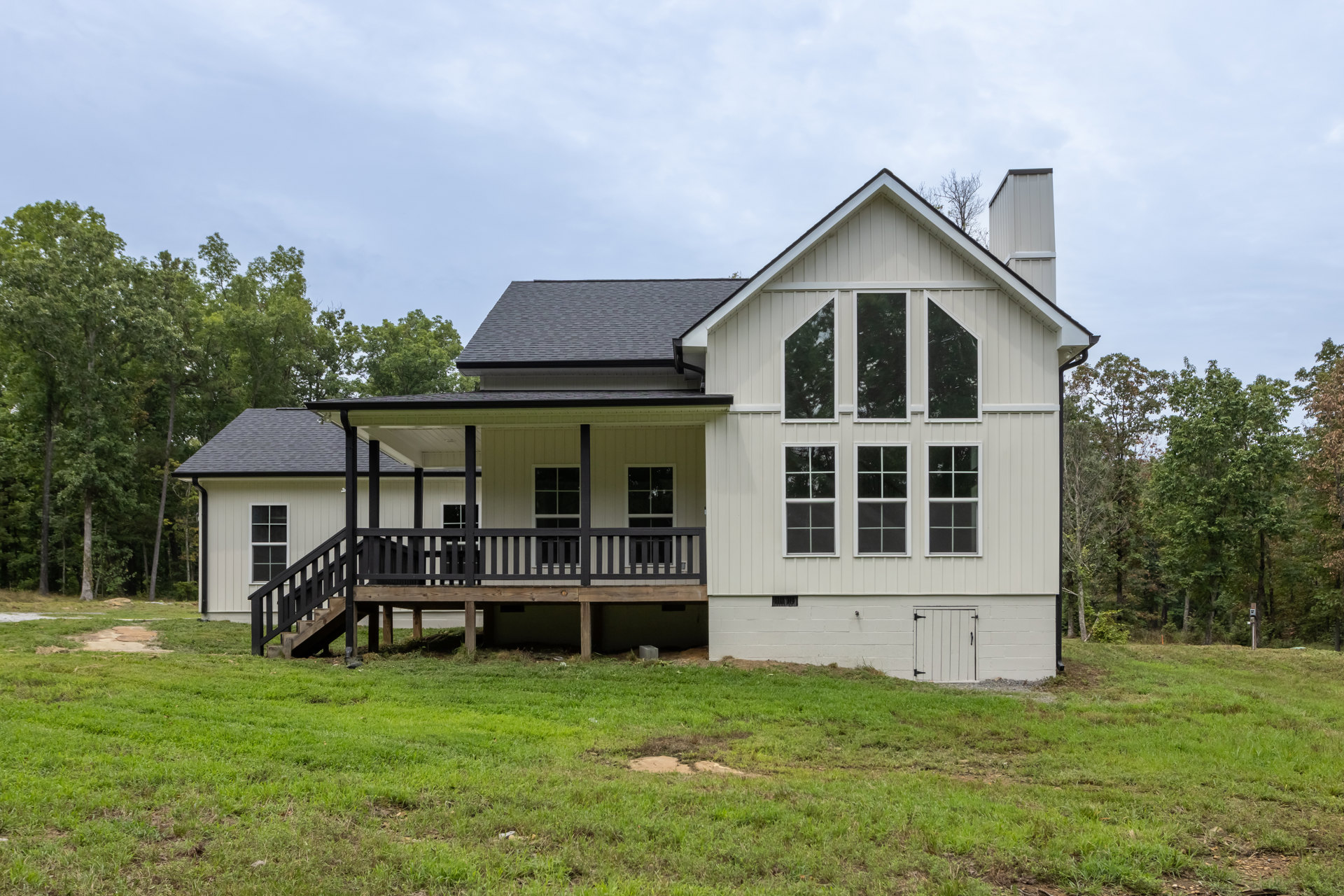 Two-story house with multiple windows, covered front porch, wooden deck with railing, black plastic wrap on one window, surrounded by grass field under partly cloudy sky