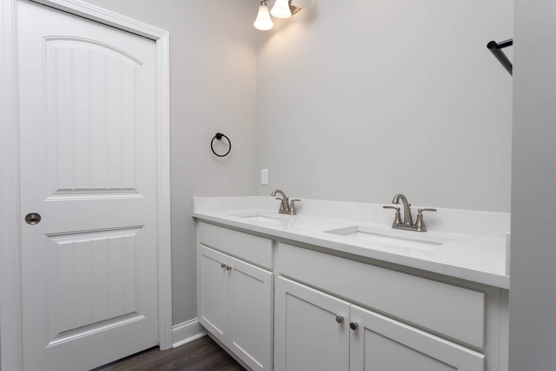 White bathroom featuring shaker-style cabinets, quartz countertops, silver hardware, white paneled door, black towel ring mounted on white wall, and recessed lighting fixture.