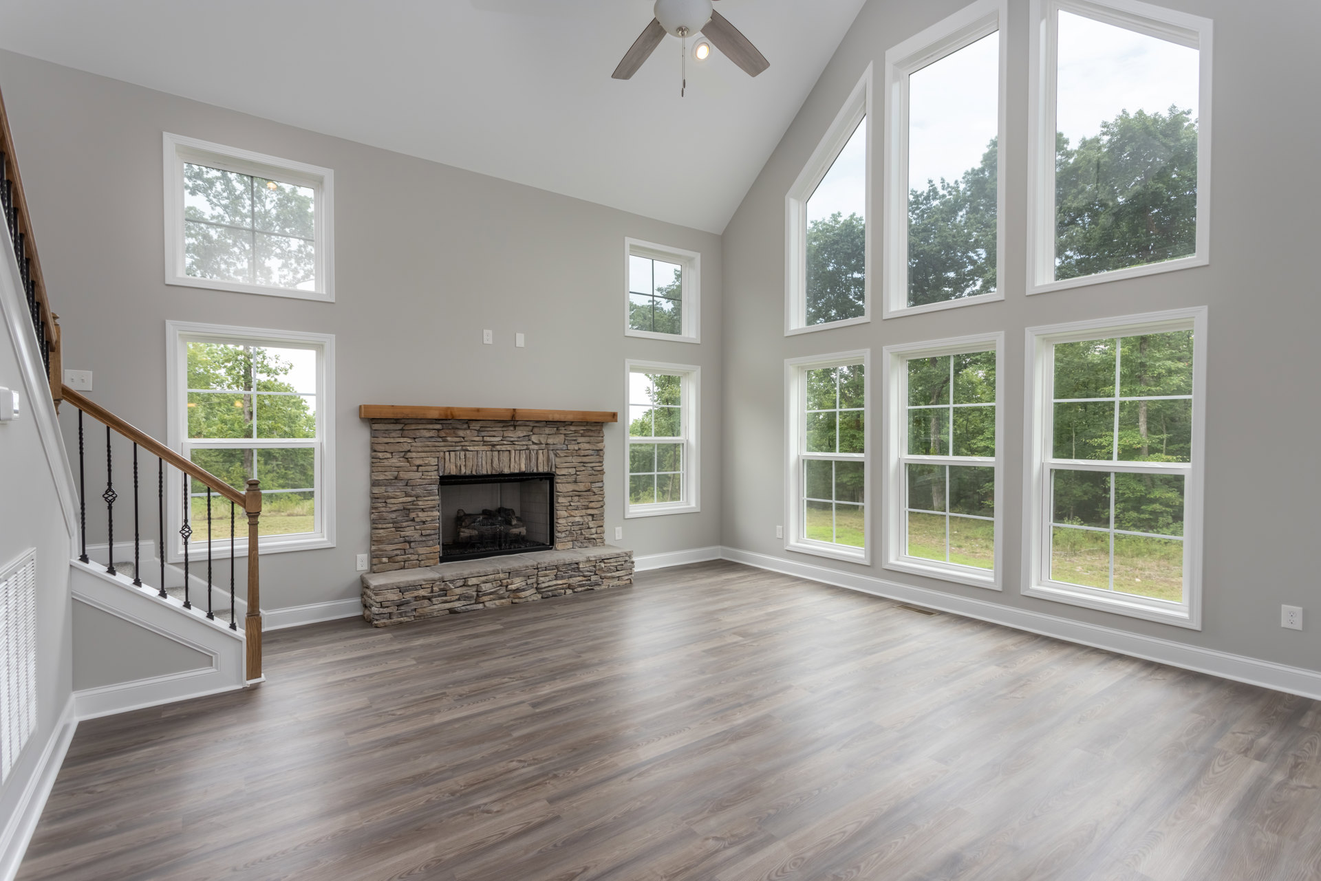 Living room with wood flooring, central fireplace featuring wire mesh screen, ceiling fan with light fixture, large window overlooking trees, and staircase with wooden railing.