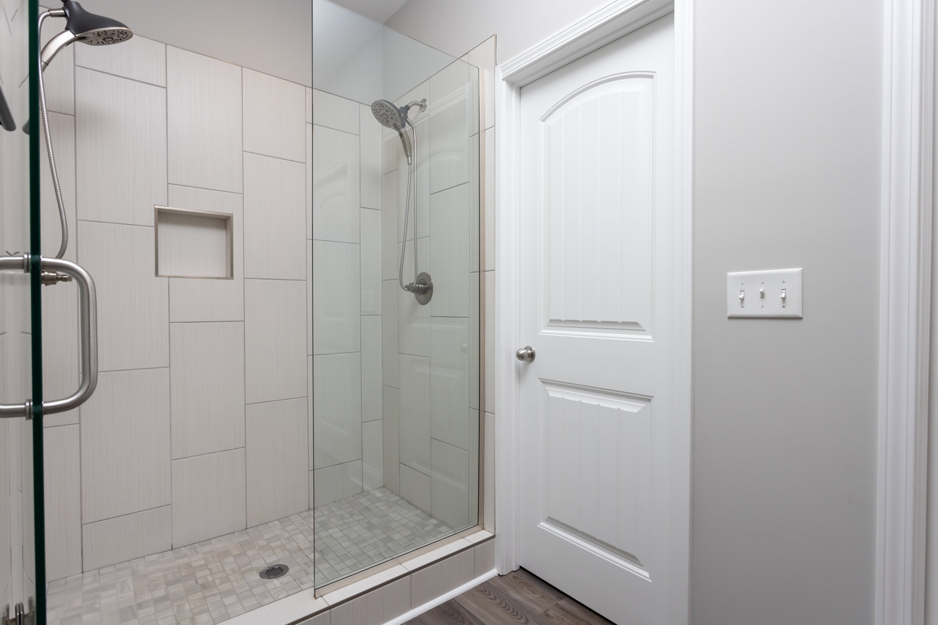 Walk-in shower with clear glass doors, chrome shower head and hose, white tile walls, row of wall switches, and modern door handle