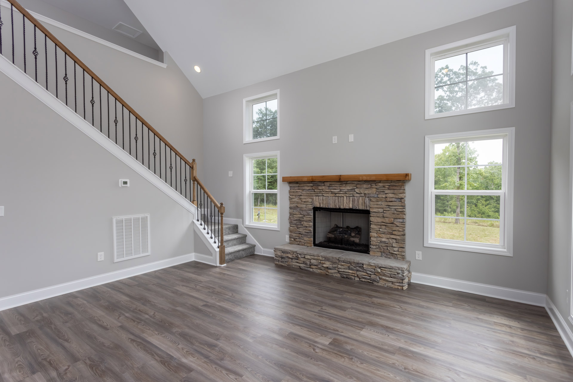 Living room with hardwood floors, wood-burning fireplace, staircase, large window showing trees, and white wall vent