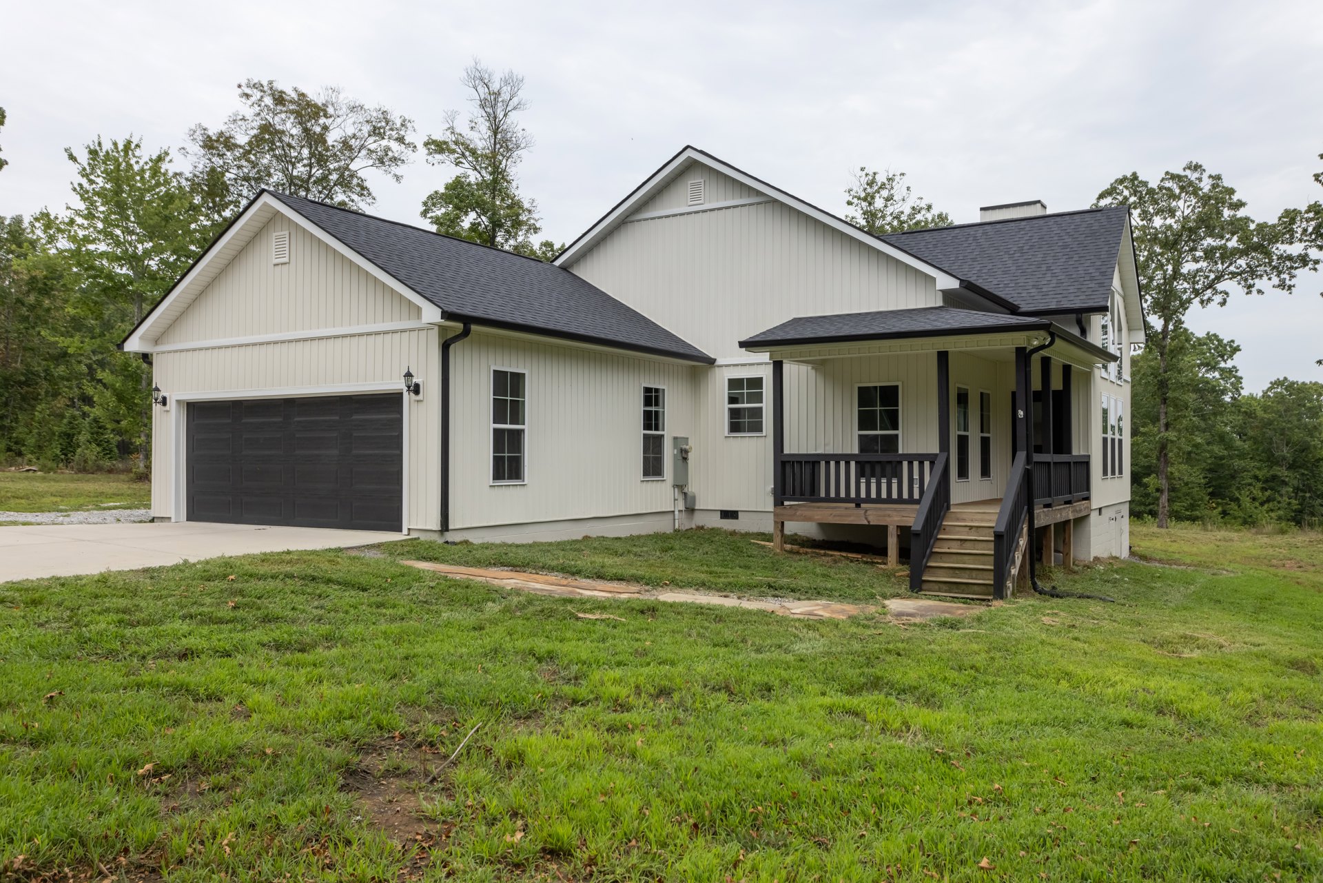 Two-story house with white siding, black-trimmed windows, covered wooden porch, wooden stairs, black garage door with white trim, and grass lawn with stone path
