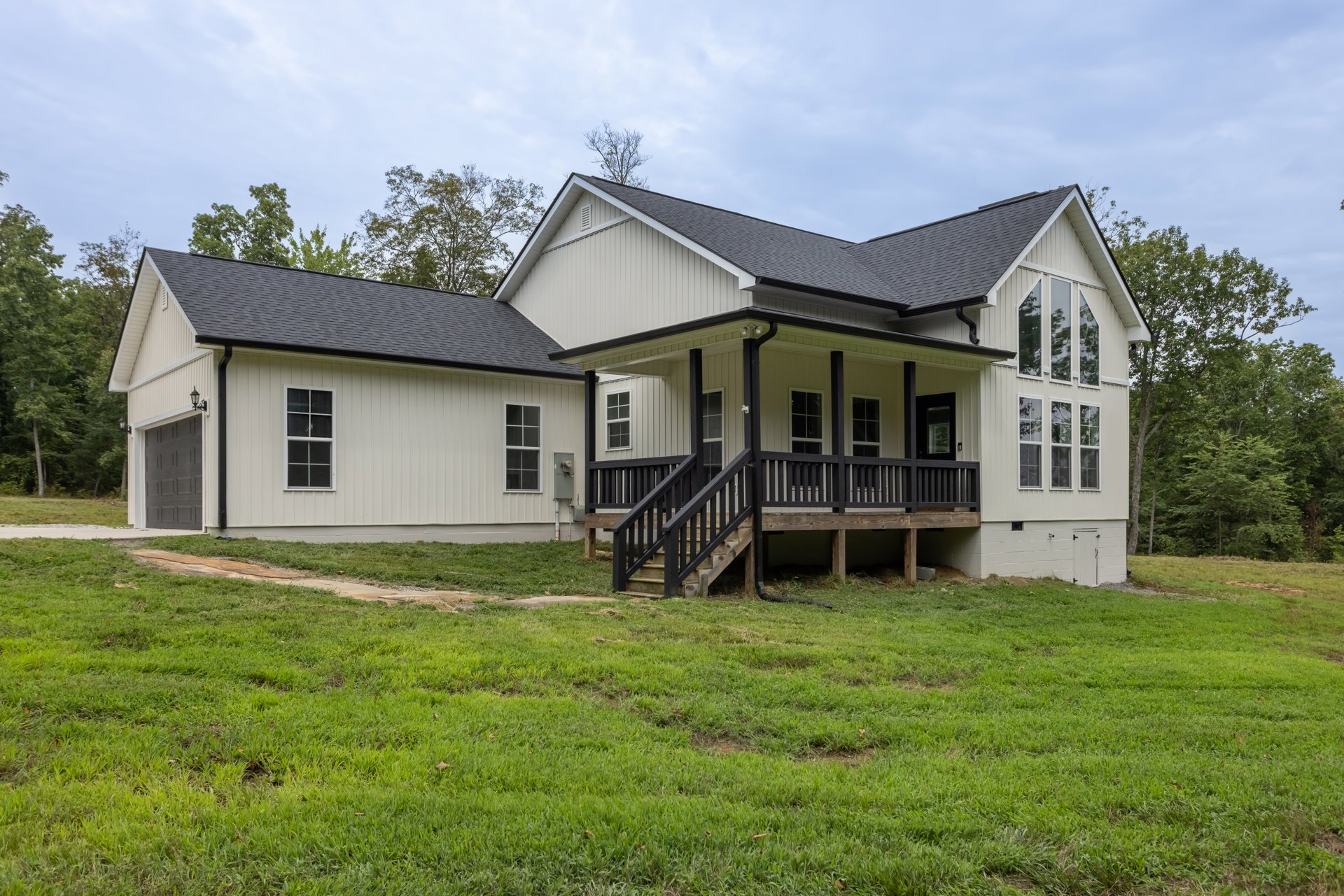 Two-story house with white siding, covered front porch, wooden stairs, large windows including one with broken glass, green grass lawn, wooden deck, and mature trees in the