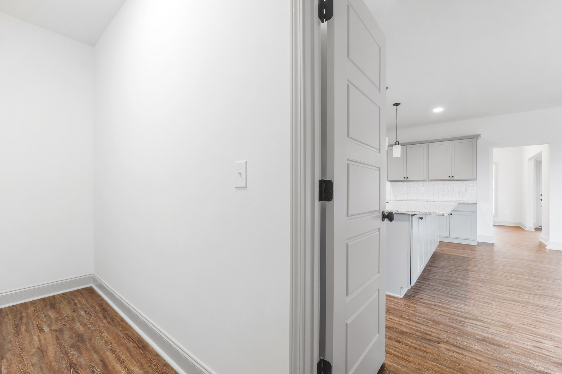 White paneled door with black knob set in a room featuring wood laminate flooring, white baseboards, white cabinets, and a light switch on the plaster wall.