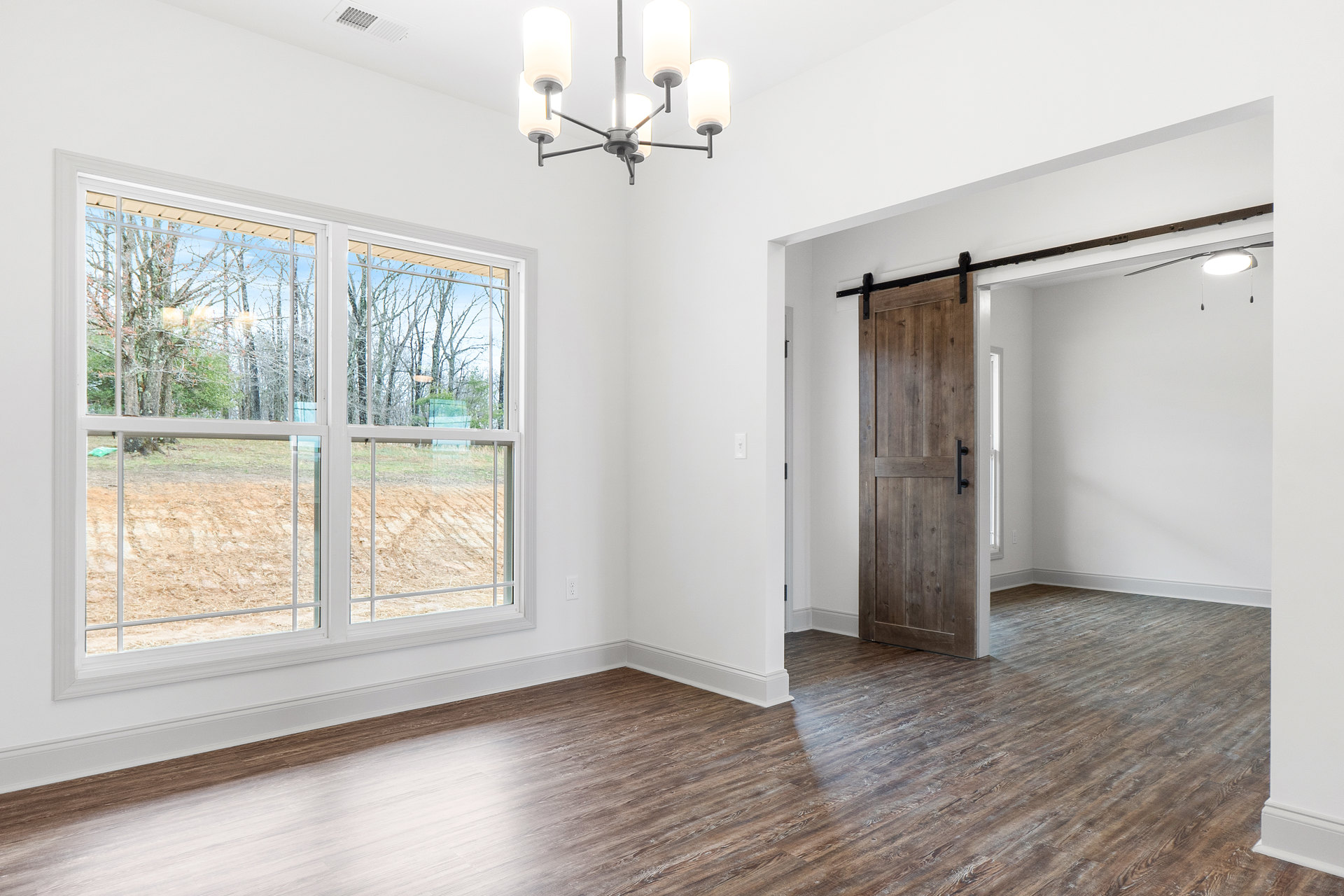 Open room with light wood flooring, large sliding glass door, window overlooking green field and trees, modern chandelier with white lights, wooden door featuring black handle