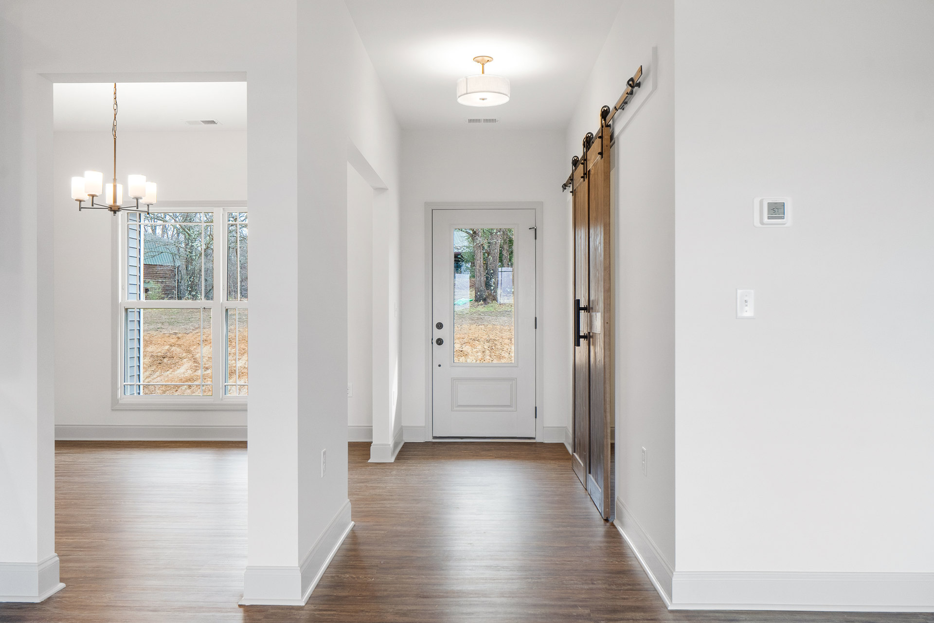 Hallway with light wood flooring, white walls, a white door with glass pane, digital thermostat mounted near door, window overlooking house and trees, modern white light fixture