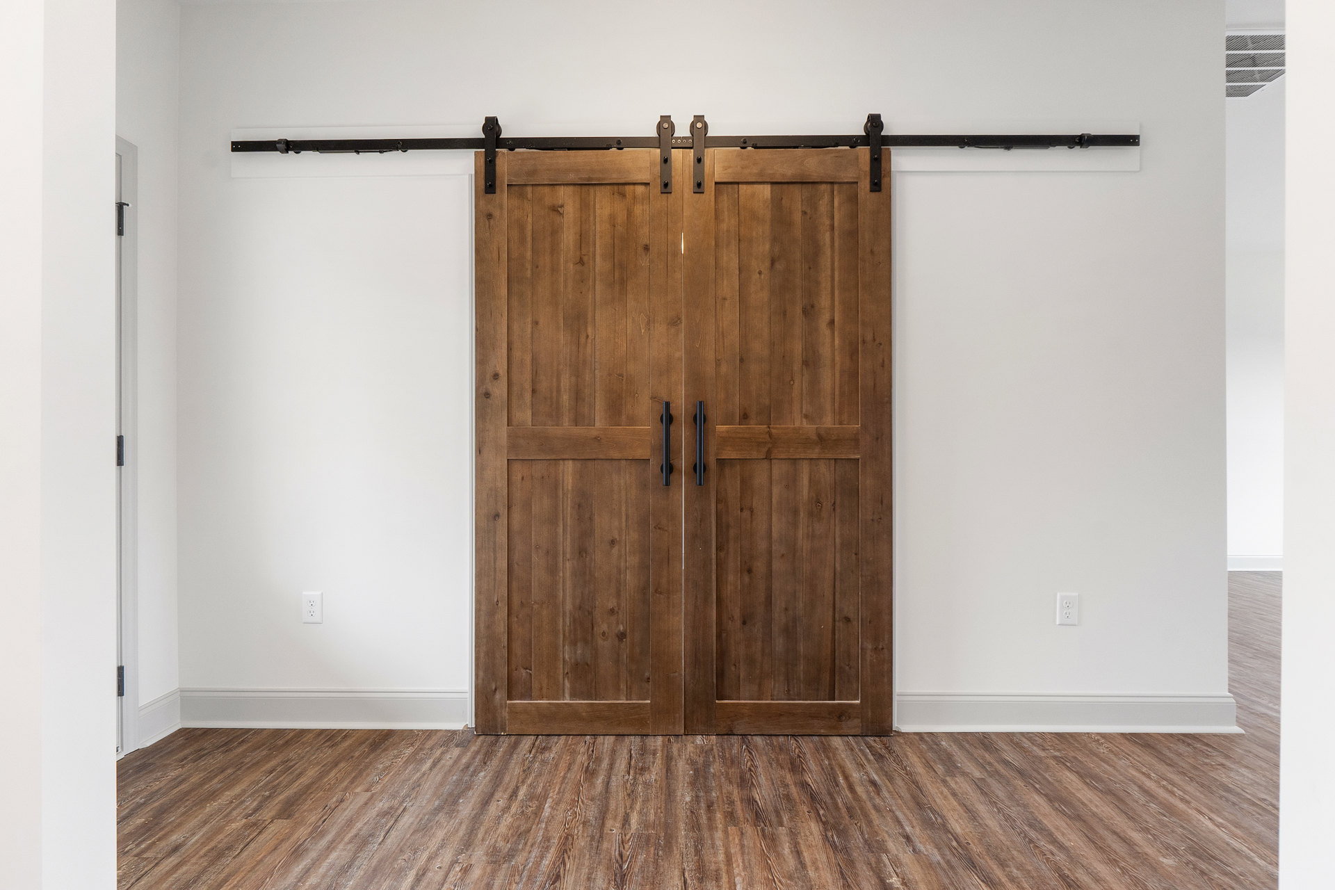Double wooden sliding doors with black handles set in a room with light hardwood plank flooring and white walls, close-up showing door details and floor vent