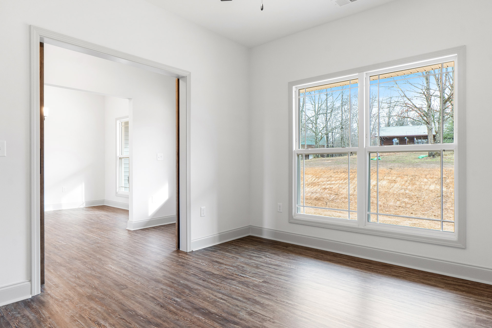 Wood flooring with white columns, large window overlooking a field and distant building, trees visible outside, plaster walls and ceiling.