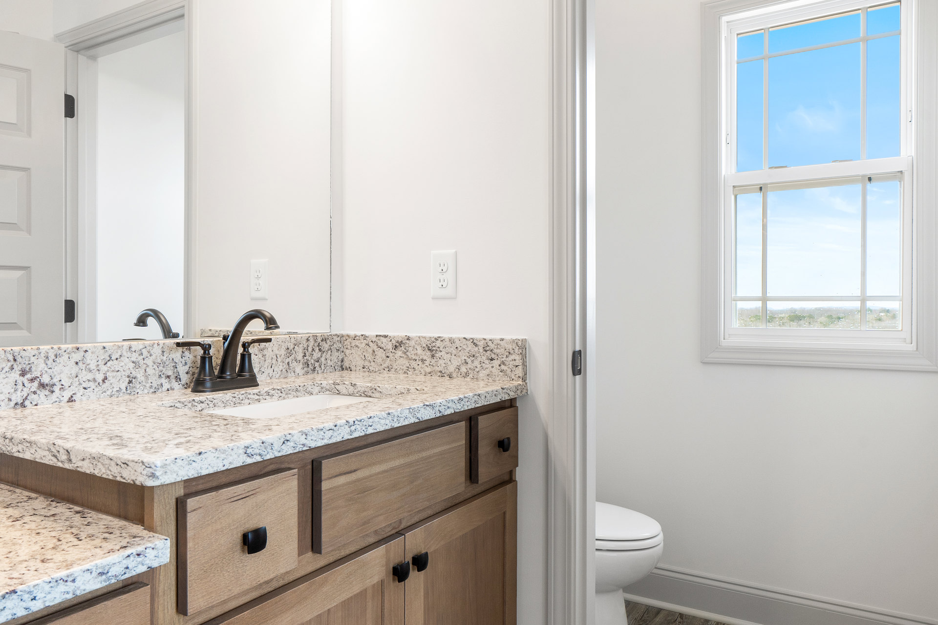 Bathroom featuring marble countertop with black faucet, white sink, closed toilet, white electrical outlet, window showing blue sky, and white cabinetry with drawers.